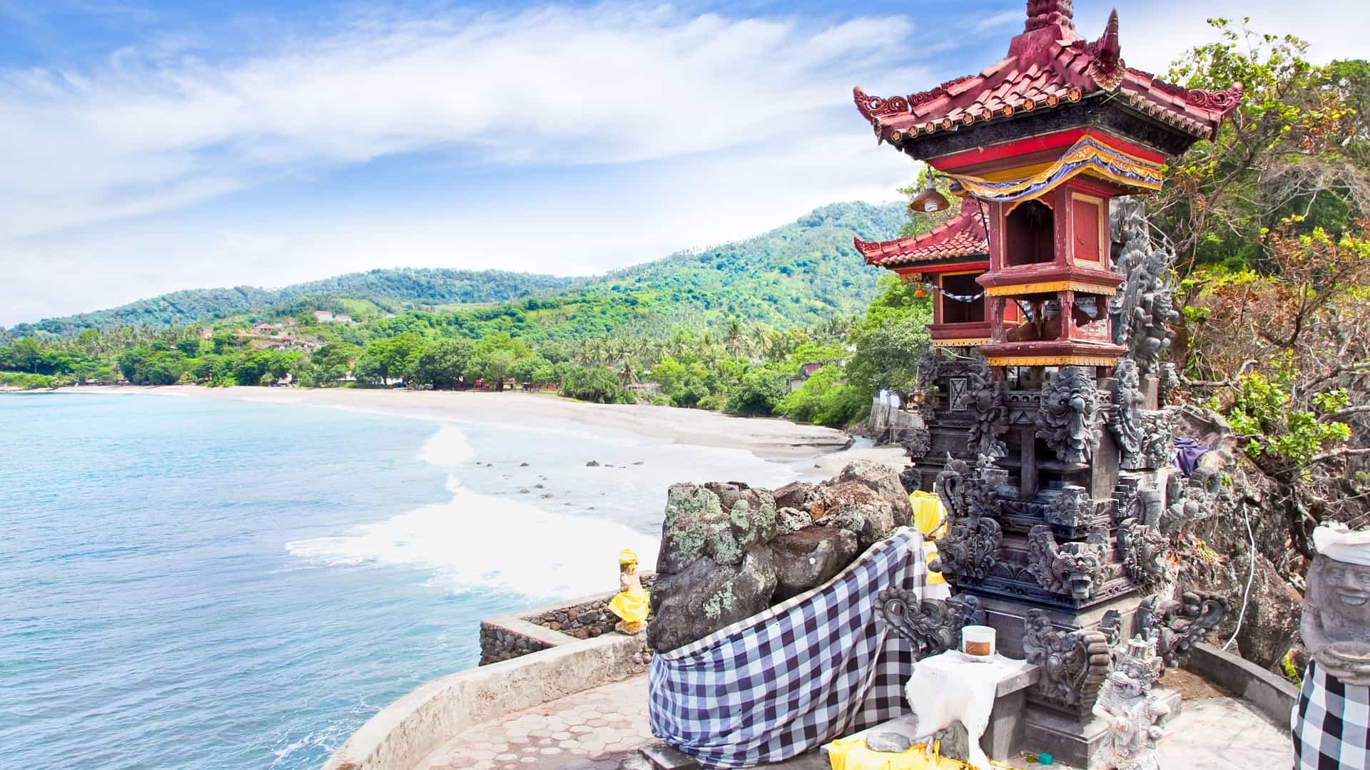 A wide shot of a traditional Balinese temple on a cliff overlooking a beautiful beach with white sand and turquoise water on the tropical island of Lombok, Indonesia.