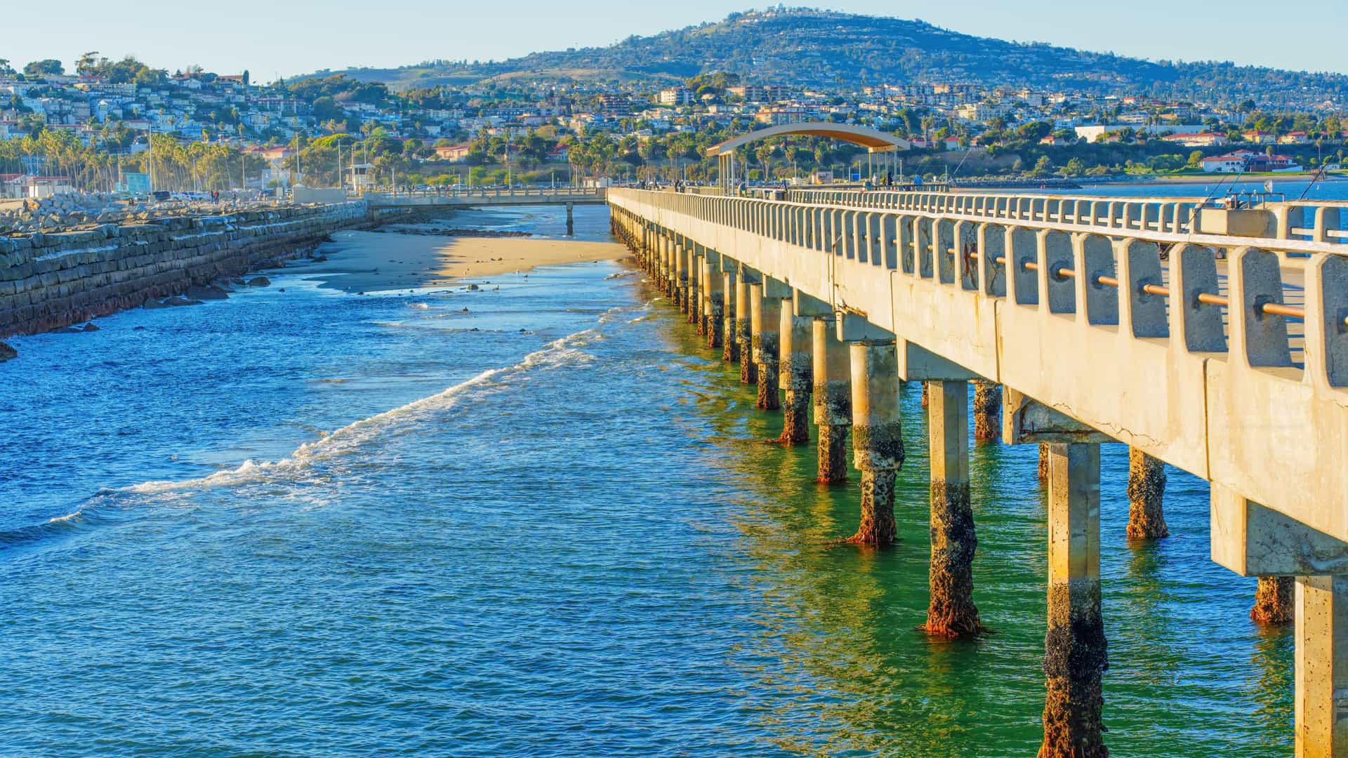 A coastal scene in Los Angeles / San Pedro featuring a long concrete pier supported by weathered columns over blue‑green water, with a rocky breakwater, sandy beach, and hillside homes and palm trees rising in the background.