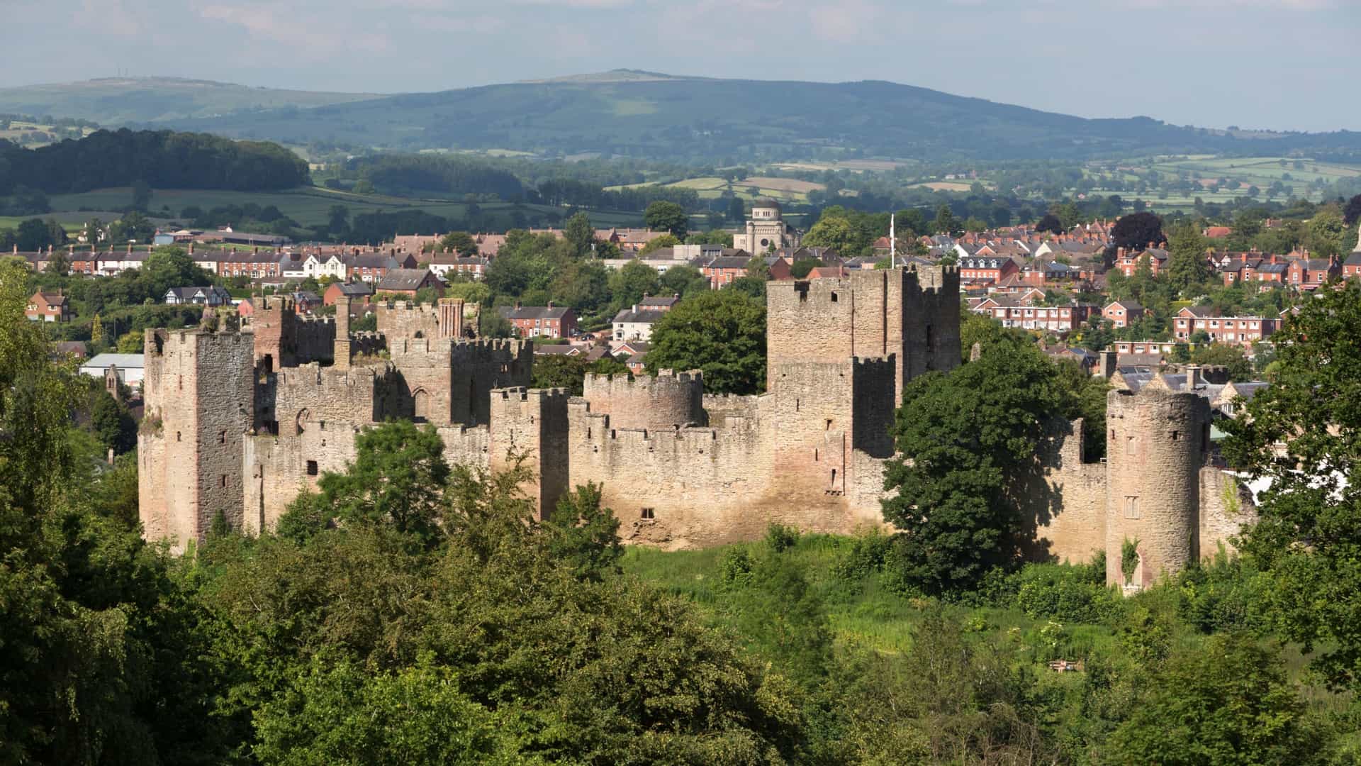 A scenic view of the medieval Ludlow Castle ruins with its stone walls and towers, overlooking the charming town of Ludlow and the rolling Shropshire hills.