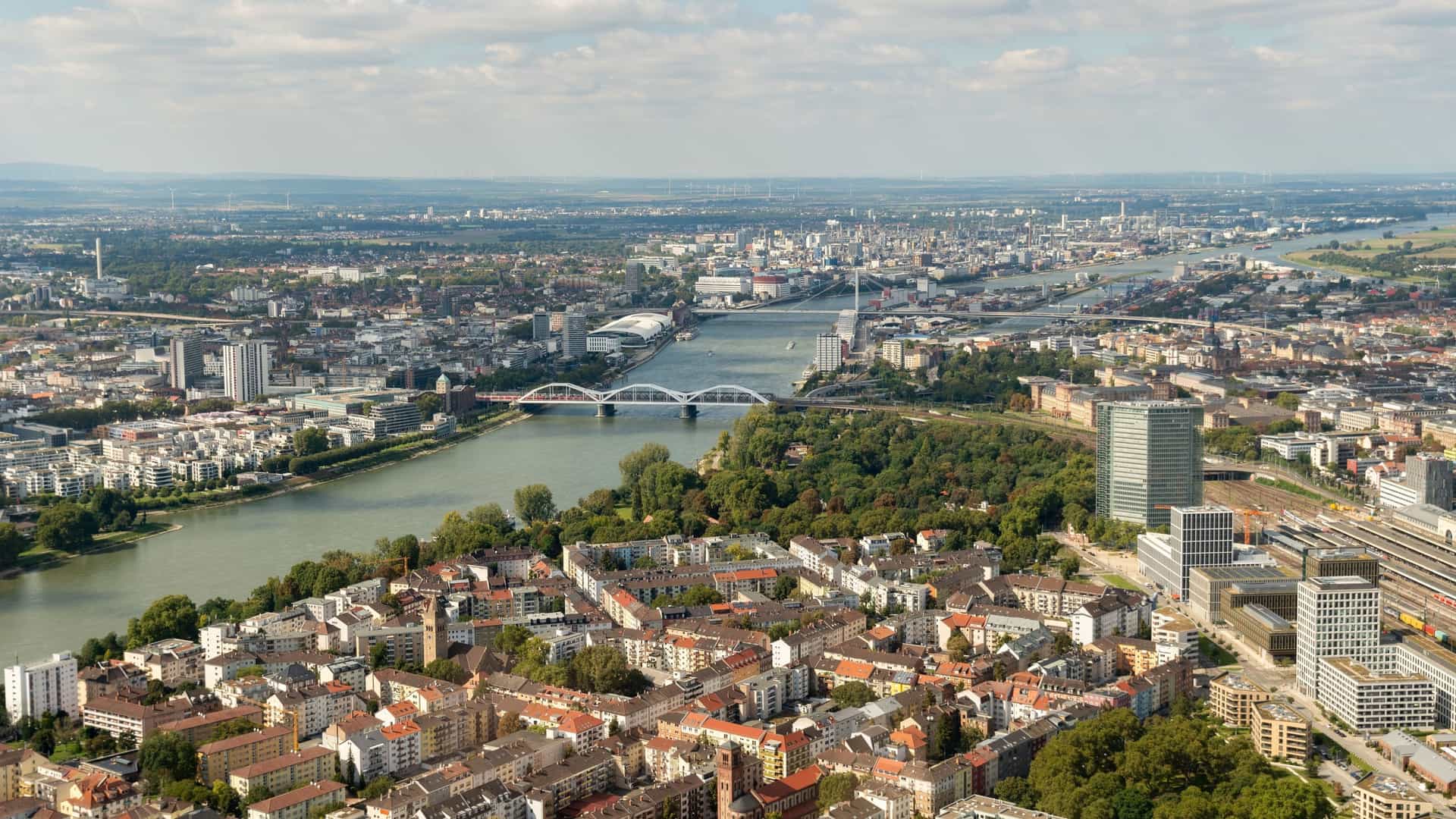An aerial panoramic shot of Ludwigshafen and Mannheim in Germany, showcasing the Rhine River flowing through the cities, with bridges and modern high-rises.