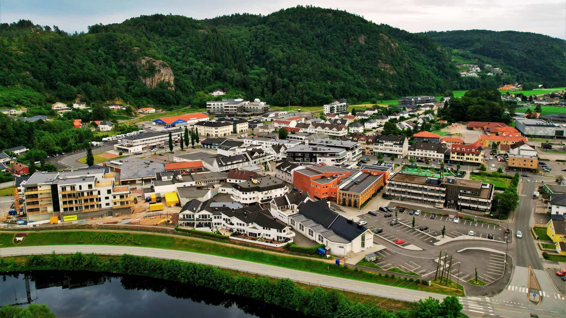 A wide, elevated landscape view of the Lyngdal town center in Southern Norway. The modern commercial buildings and infrastructure of the town are situated in a valley, tightly surrounded by steep, lush green hills and forests under a bright sky. The town is concentrated around the main road and commercial strip.