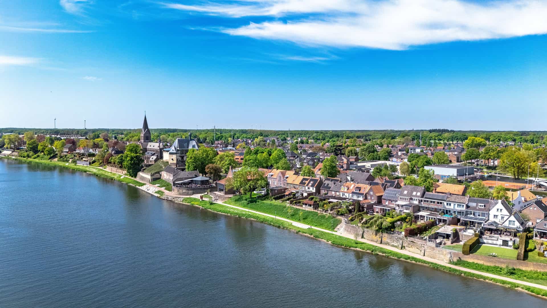 A peaceful riverside scene in Maas / Meuse River with traditional homes and a tall‑steepled church surrounded by lush greenery, a walking path along calm water, and distant forests and wind turbines under a clear blue sky.
