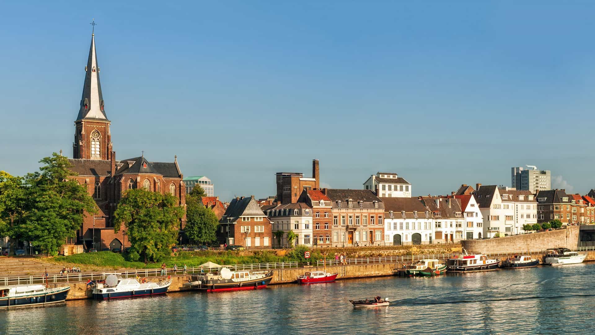 A scenic view of Maastricht, Netherlands, with the historic St. Servatius Church and traditional buildings lining the Maas River, with boats docked along the waterfront.