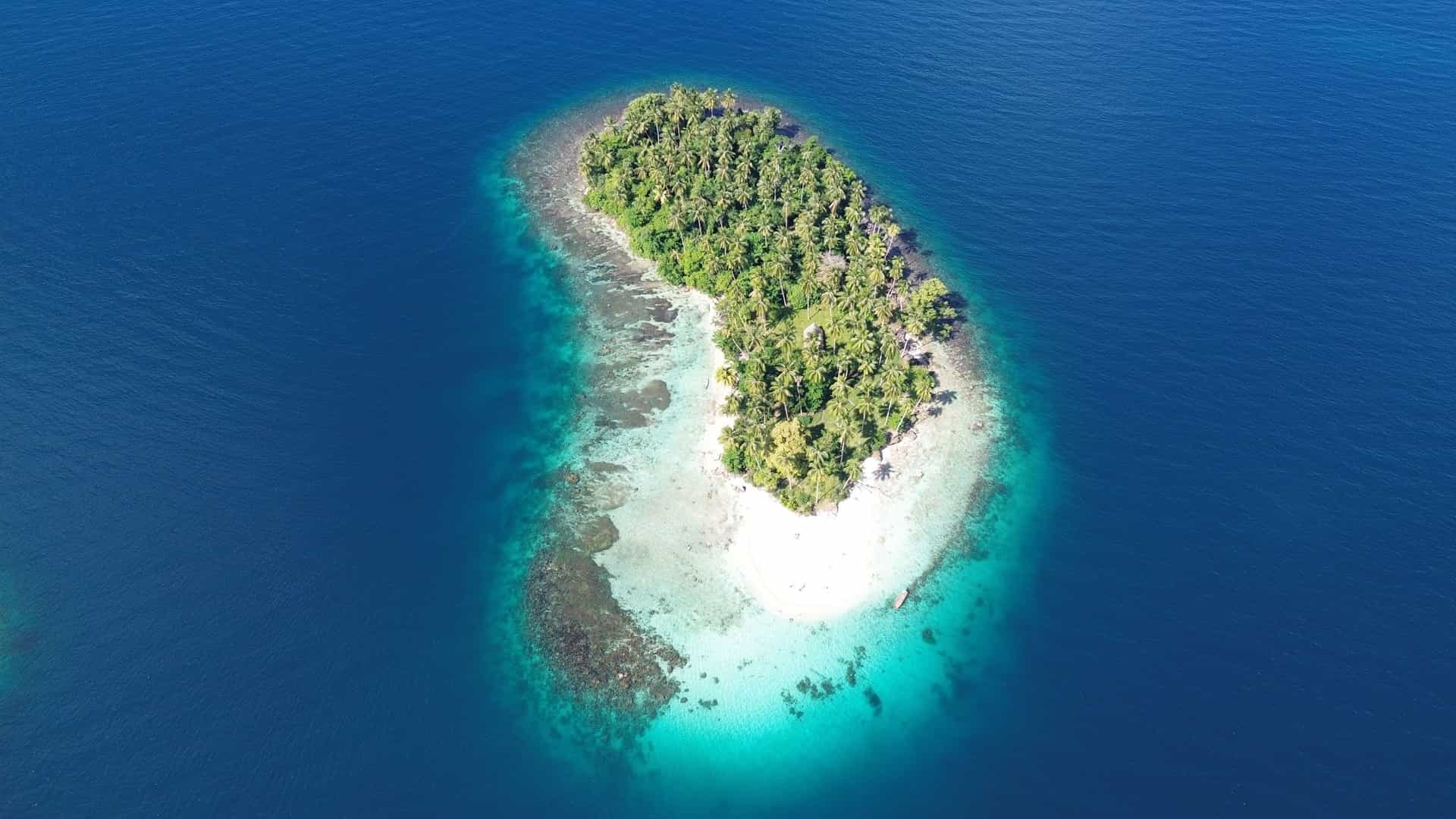 An aerial shot of a small, uninhabited tropical island near Madang, Papua New Guinea, with a lush green interior and a white sand beach surrounded by vibrant turquoise water.