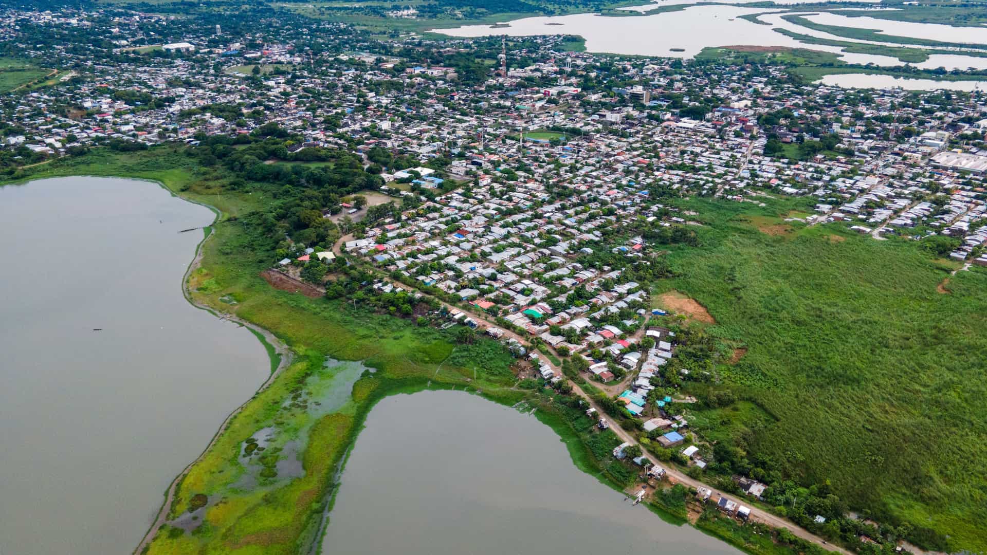 Aerial view of Magangué, Colombia, a densely built town with colorful roofs, bordered by a murky river/lake and lush green land, under an overcast sky.
