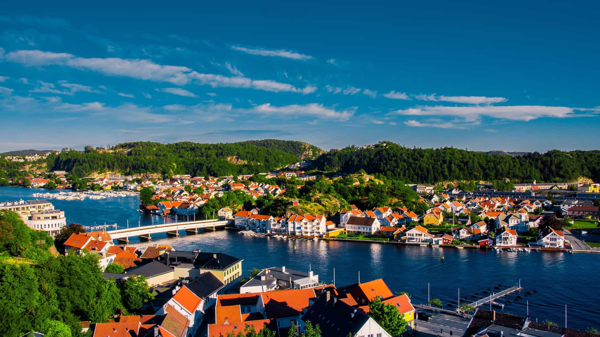 A scenic aerial shot of the coastal town of Mandal, Norway, with colorful houses clustered along the water and a bridge connecting two sides of the harbor.