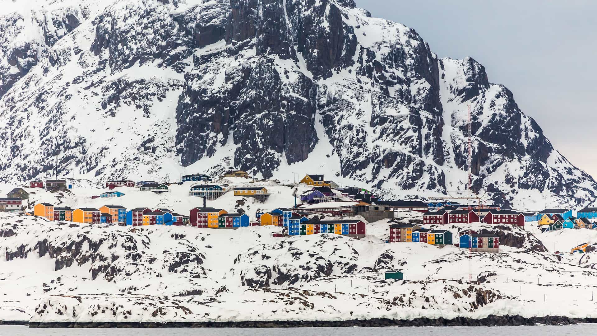 A stunning winter landscape of colorful houses nestled at the base of a massive snow-covered mountain in the remote town of Maniitsoq, Greenland.