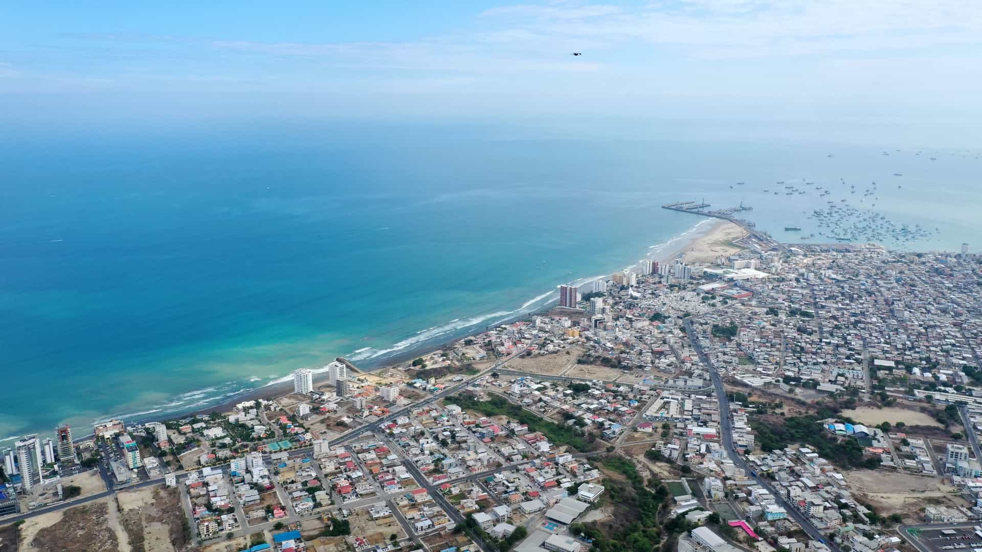 A high-altitude aerial view of a sprawling coastal city with dense housing along a turquoise ocean and a busy harbor filled with boats.