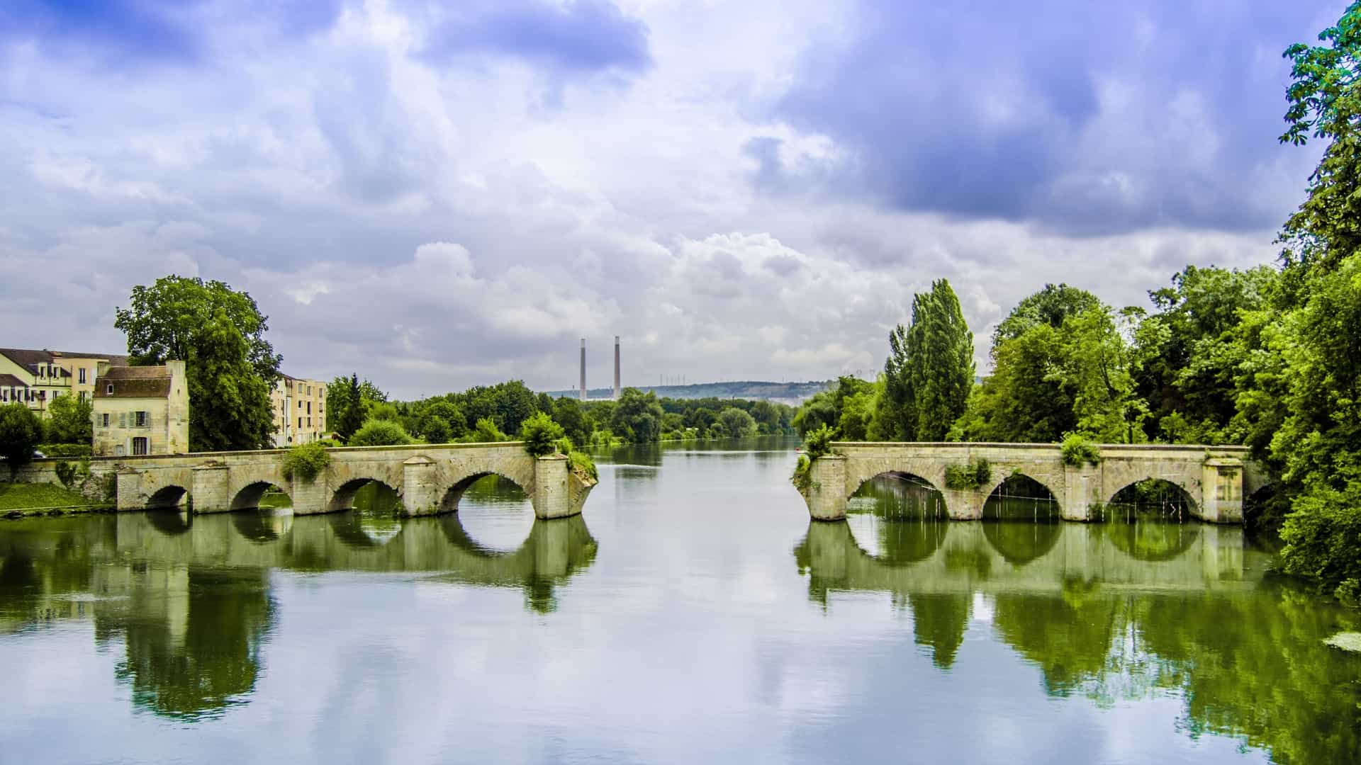 A picturesque view of the old stone bridge with multiple arches spanning the calm waters of the Seine River in Mantes-la-Jolie, France.