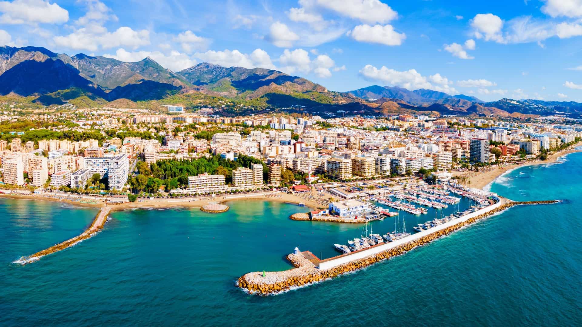 An aerial view of Marbella, Spain, shows a city, beaches, and a marina against a backdrop of steep, green mountains.