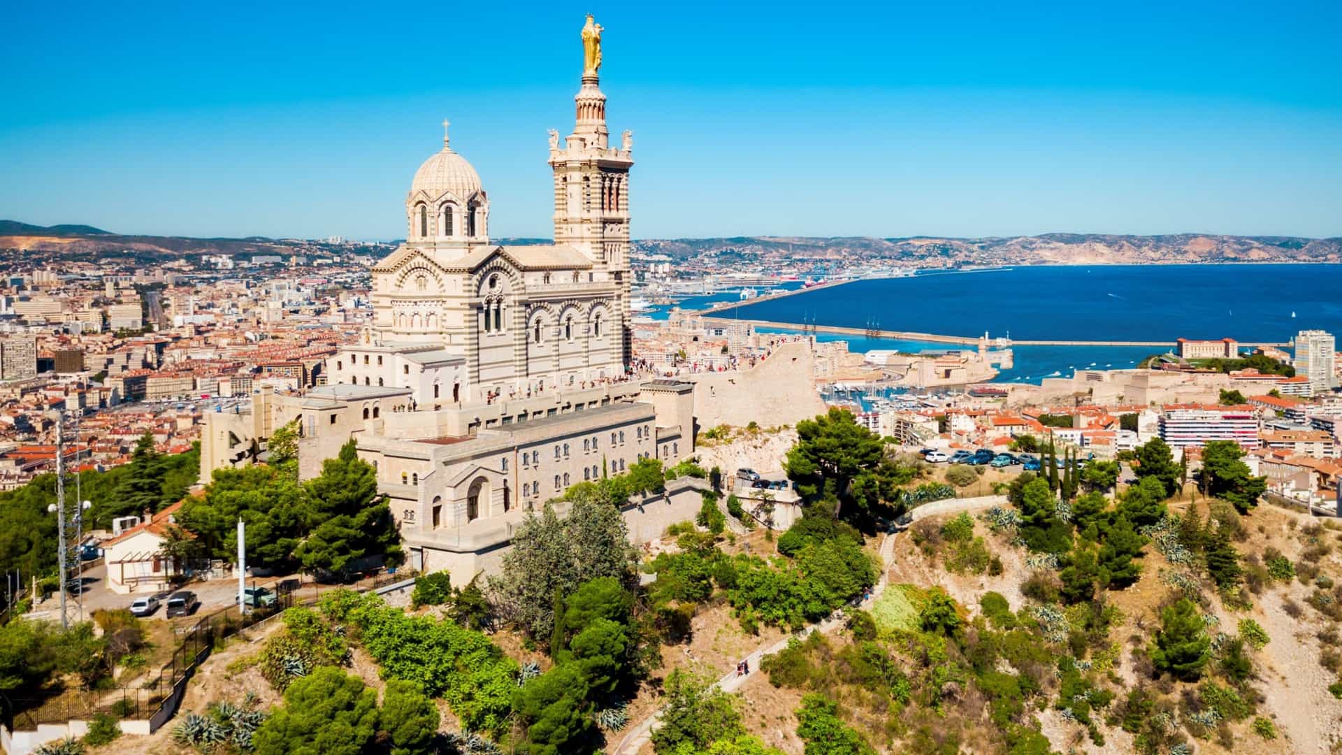 The Byzantine-style Notre-Dame de la Garde Basilica in Marseille, France, is perched on a hill, featuring alternating bands of white and green stone, and crowned by a large golden statue of the Virgin Mary and Child.