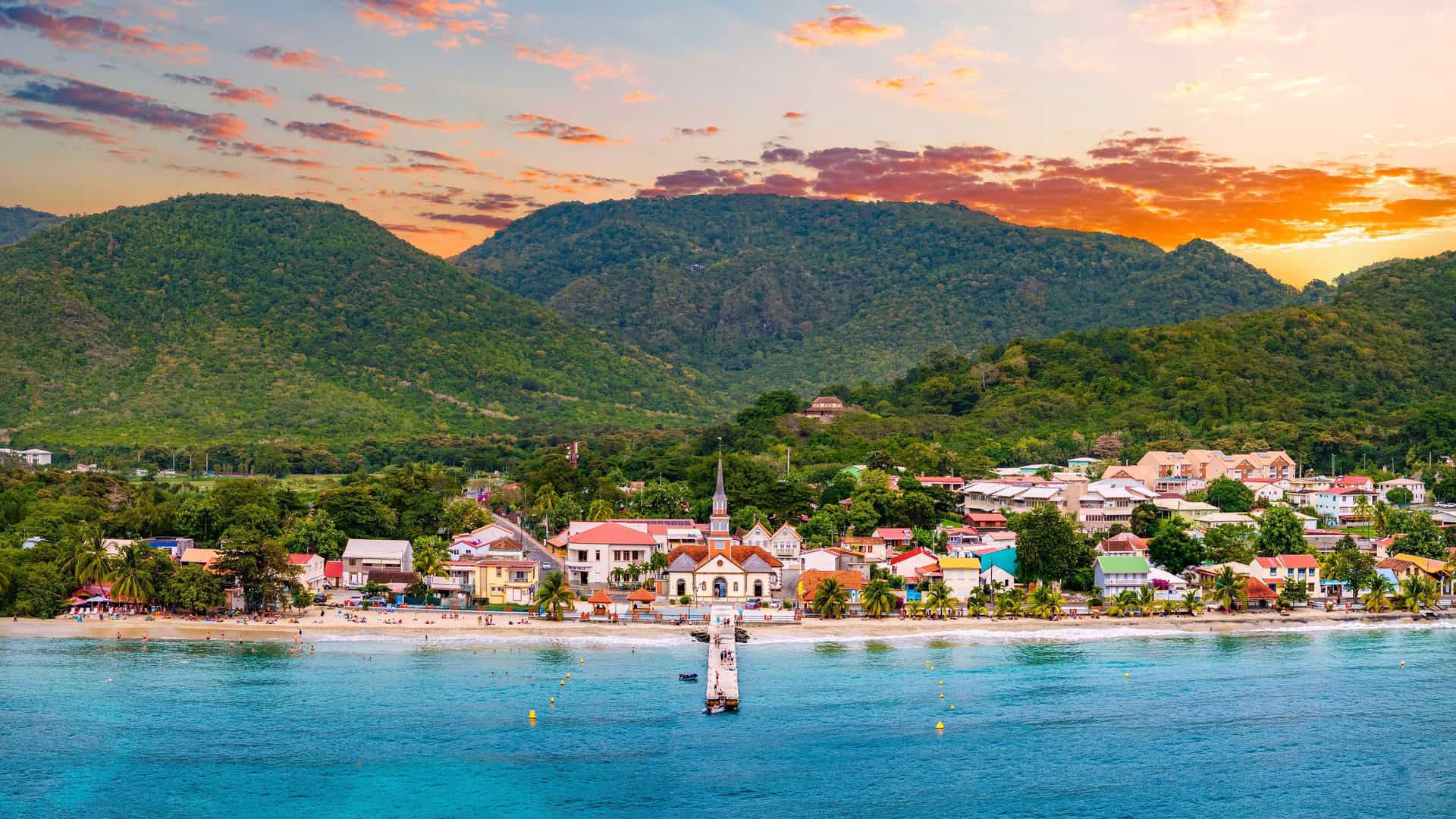 A vibrant sunset view of the coastal town of Martinique, with colorful houses and a church steeple on the beach, nestled against lush green hills.