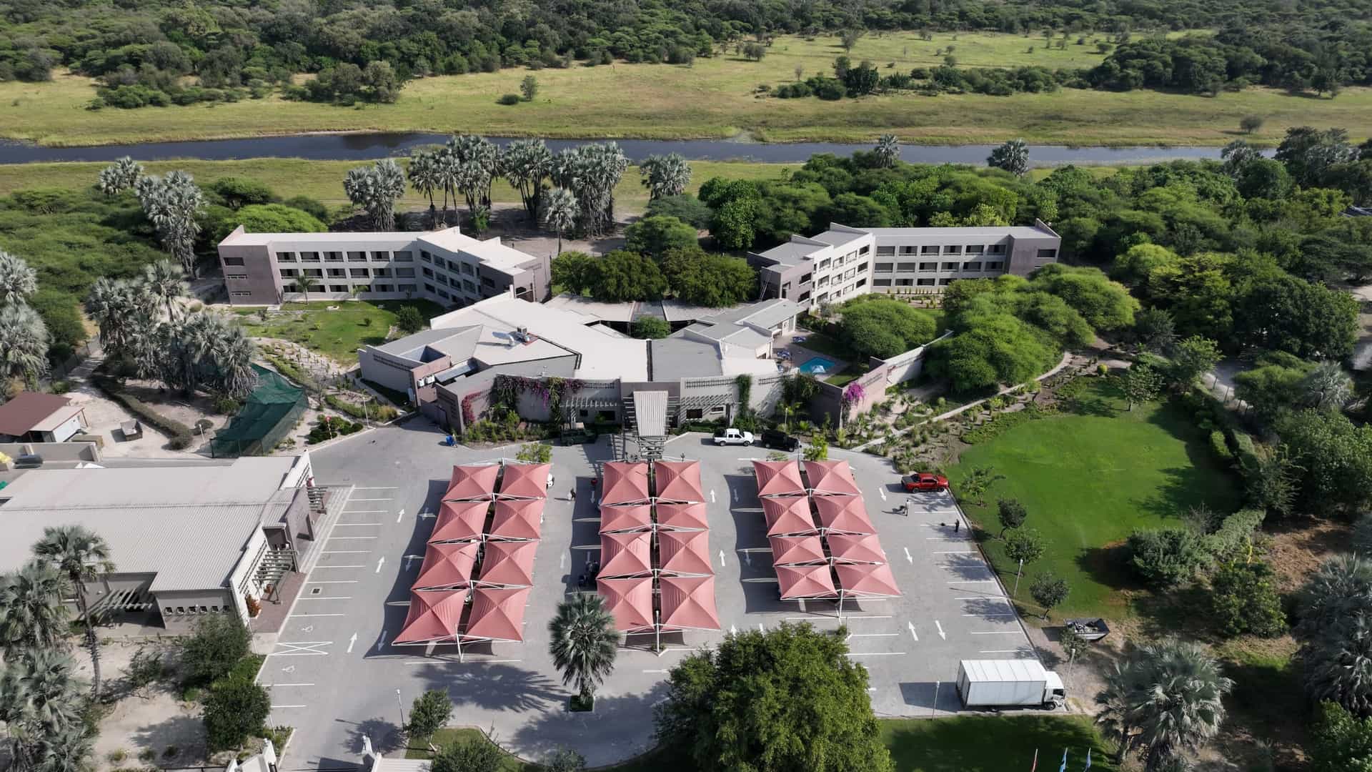 An aerial view of a resort hotel complex with pink shade structures in a parking lot, surrounded by green trees and a river.
