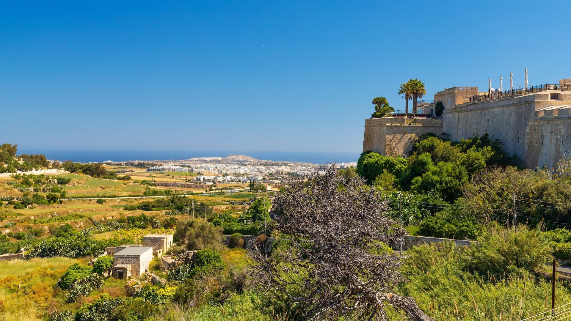 A high, fortified stone wall of the historic city of Mdina overlooks the countryside and the distant blue Mediterranean Sea in Malta.