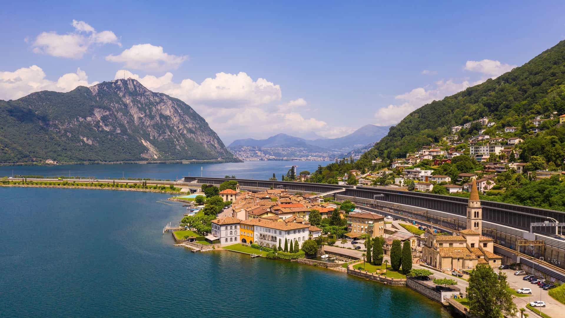 "An aerial view of the lakeside village of Melide, Switzerland, showcasing the historic buildings and a church with a bell tower on the shores of Lake Lugano with Monte San Salvatore in the background. "