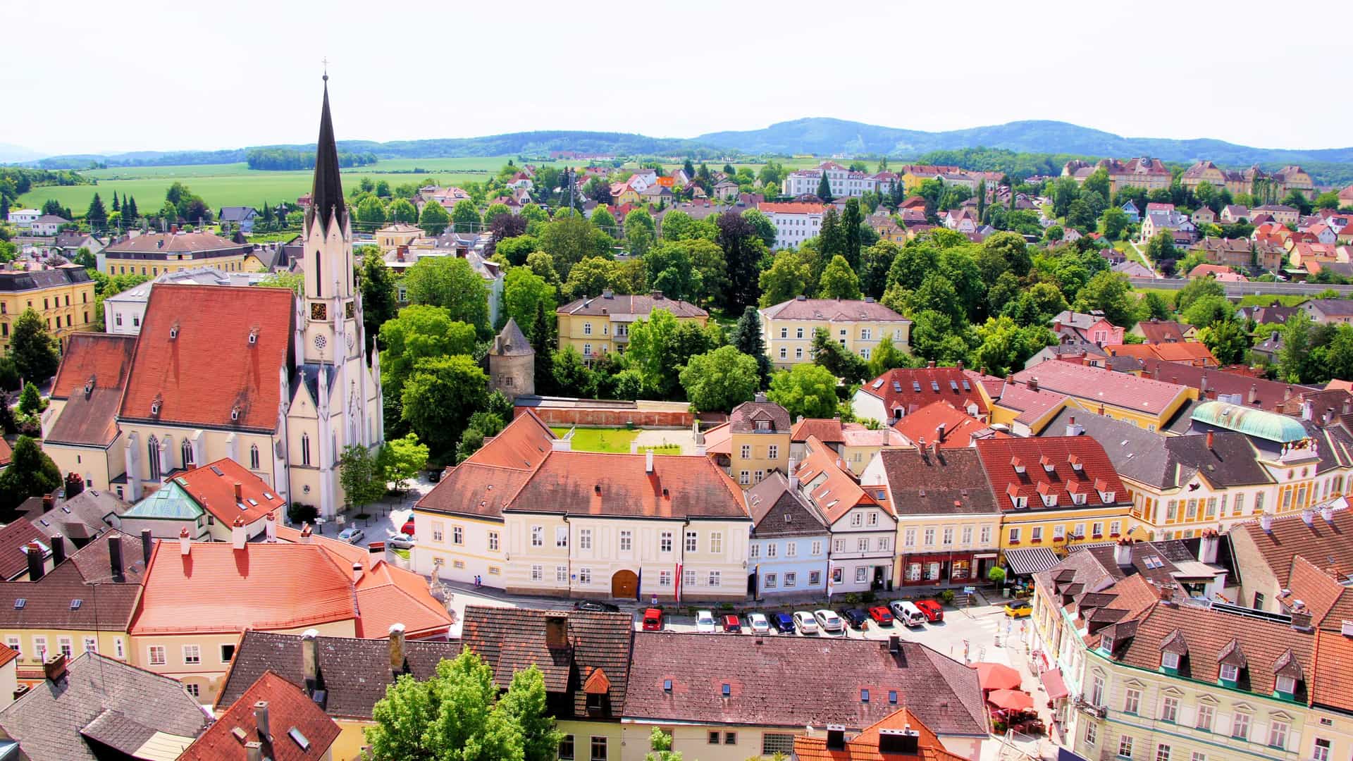 A scenic aerial view of the historic town of Melk, Austria, with a prominent church steeple and traditional red-roofed buildings surrounded by rolling green hills.