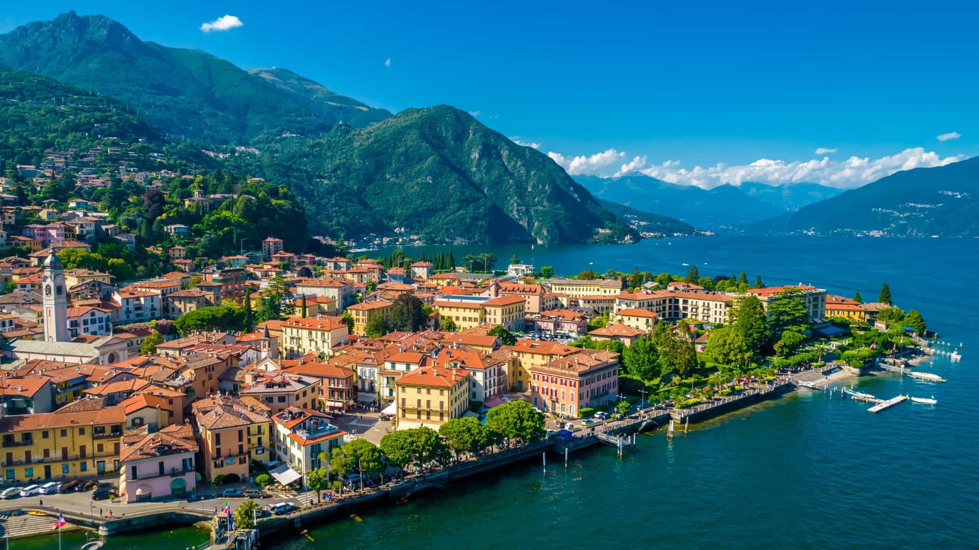 An aerial view of the lakeside town of Menaggio, Italy, with red-roofed buildings nestled against steep green mountains.