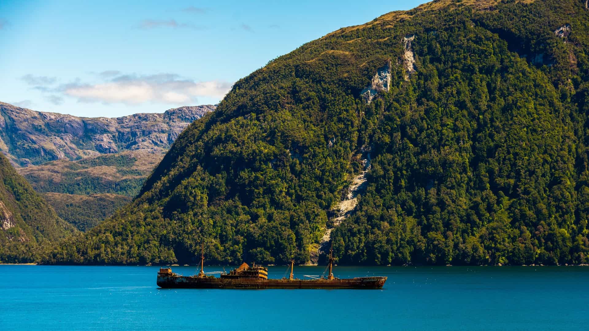 A rusty shipwreck of the cargo ship "Capitán Leonidas" rests in the calm, blue waters of Messier Channel in the Chilean Patagonian fjords, with lush green, forested mountains towering in the background.