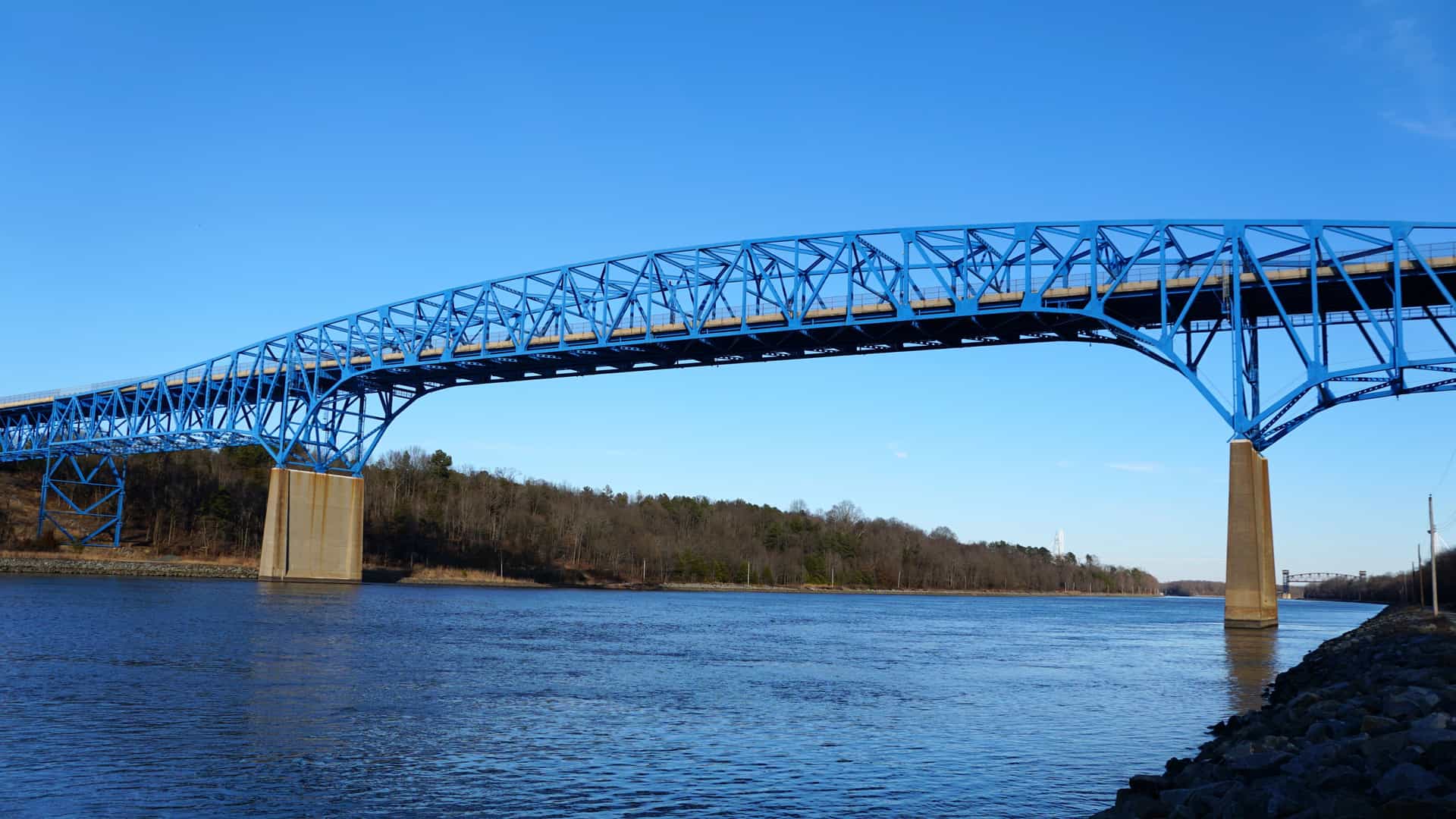 A wide river scene in Middletown featuring a large blue truss bridge supported by tall concrete piers, spanning calm water with forested banks and a clear blue sky completing the view.