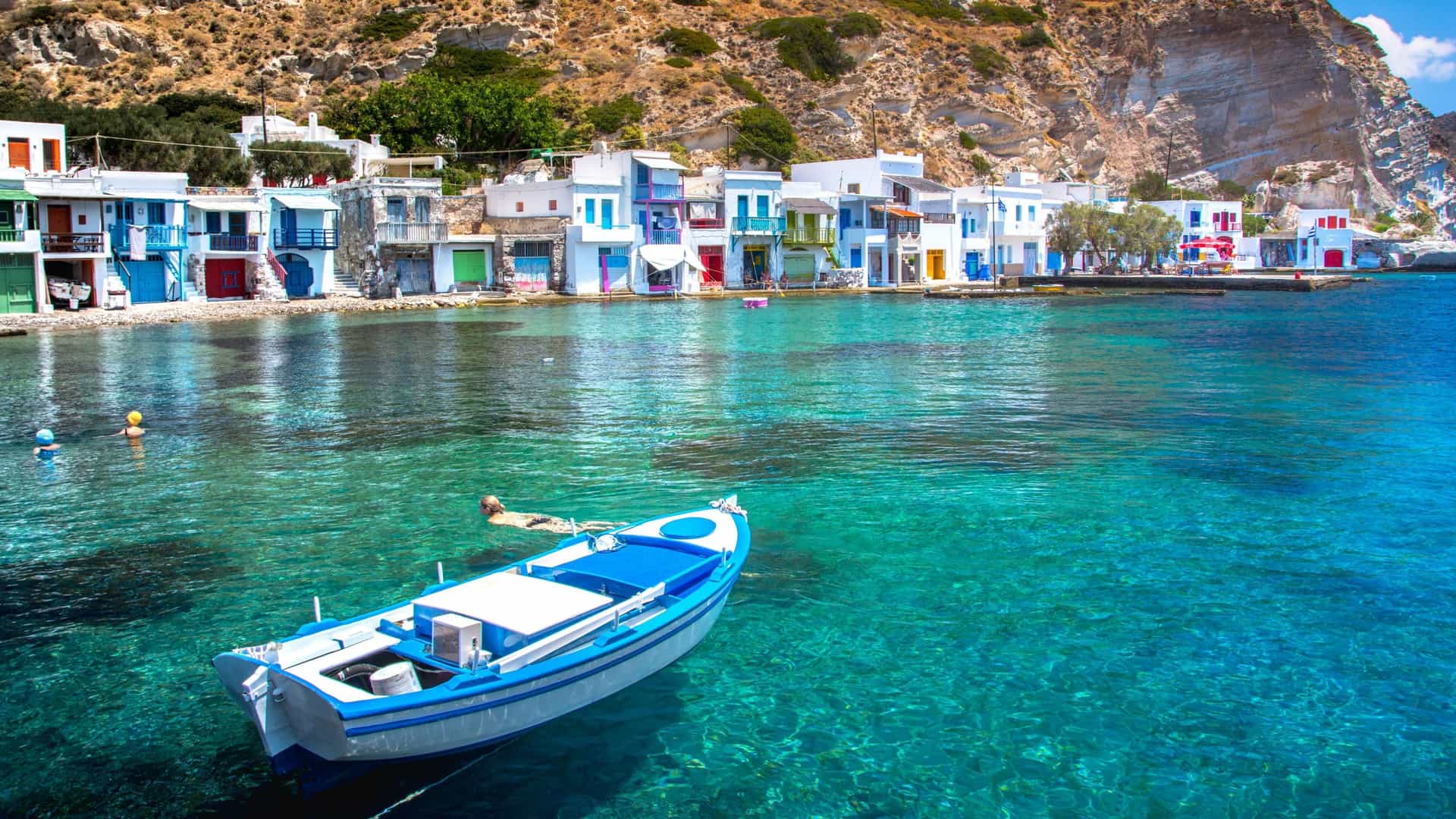 A colorful image of a traditional Greek boat moored in the turquoise water of a fishing village. The village features charming whitewashed houses with brightly painted doors, built at the base of a cliff.