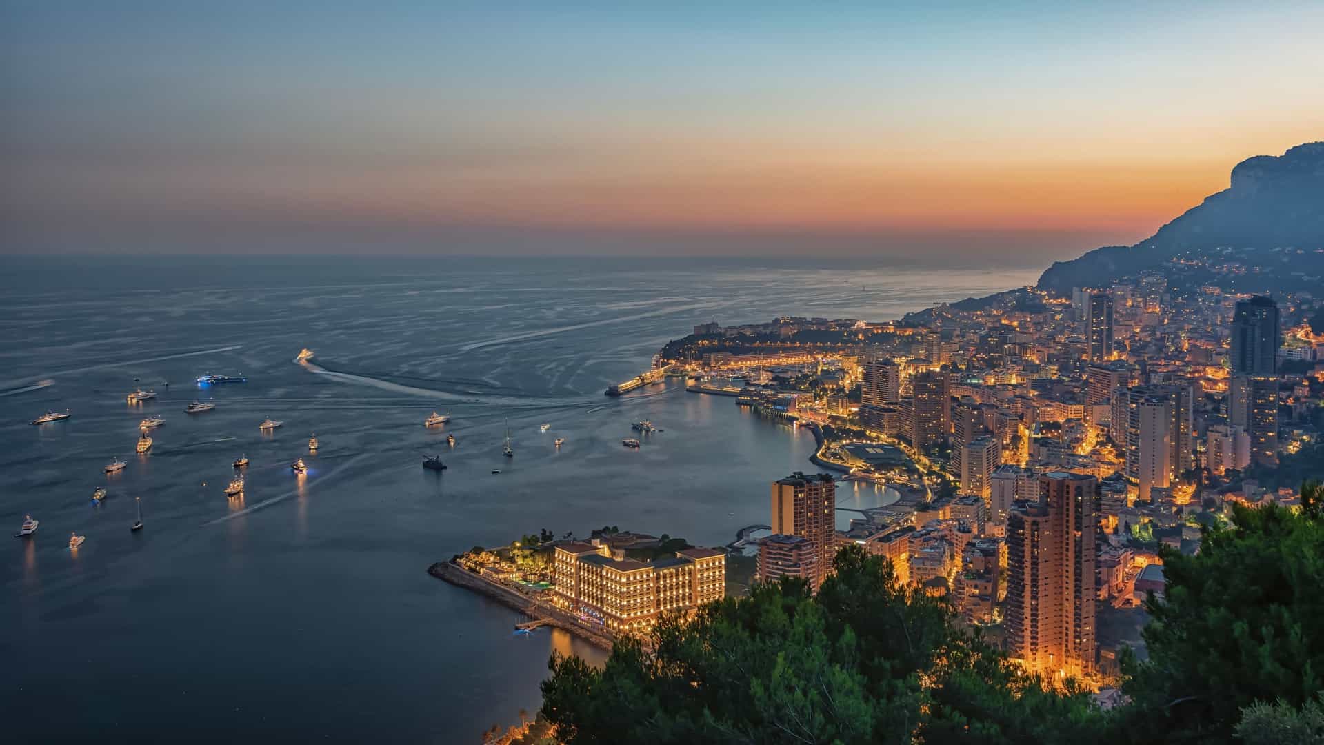 A high-angle panoramic view of the Principality of Monaco, showing the densely packed city skyline of Monte Carlo and the port (Port Hercule) filled with luxury yachts, all set against the steep, green hills of the French Riviera under a bright sky. The Prince's Palace on "The Rock" and the Mediterranean Sea are prominent.