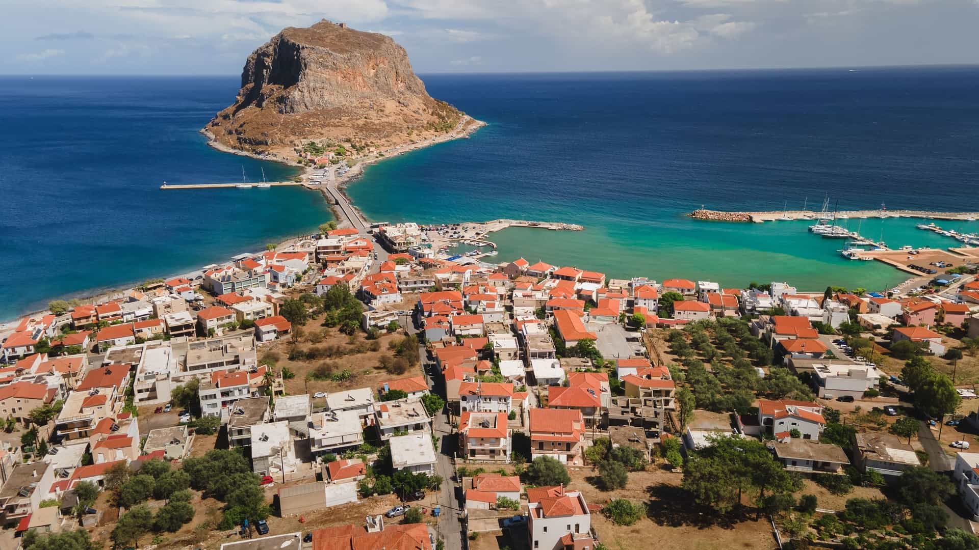 An aerial view of the historic town of Monemvasia, Greece, with its red-roofed buildings nestled on a small peninsula connected to the mainland by a single causeway.