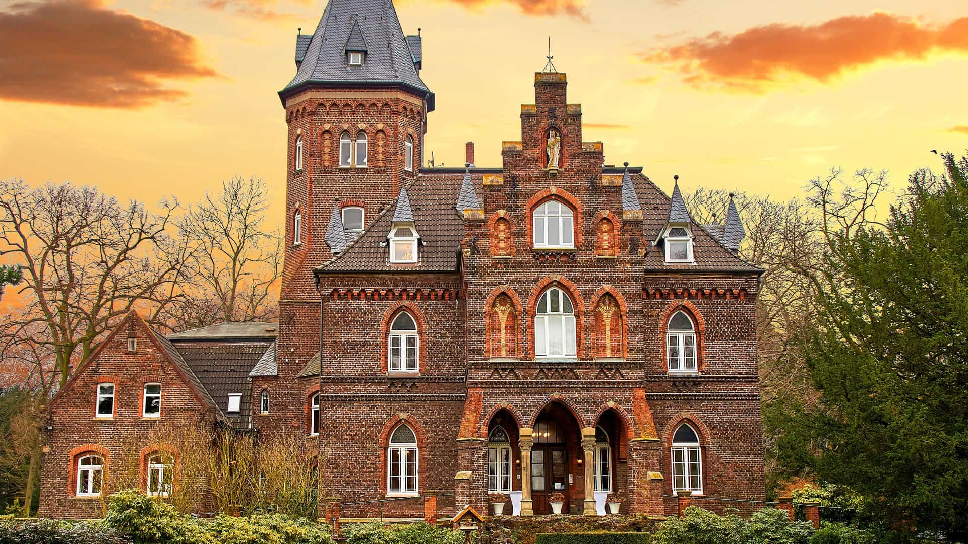 "A front view of the historic Monheim Castle in Monheim am Rhein, Germany, a red brick building with a tower, set against a stunning orange and gold sunset sky. "