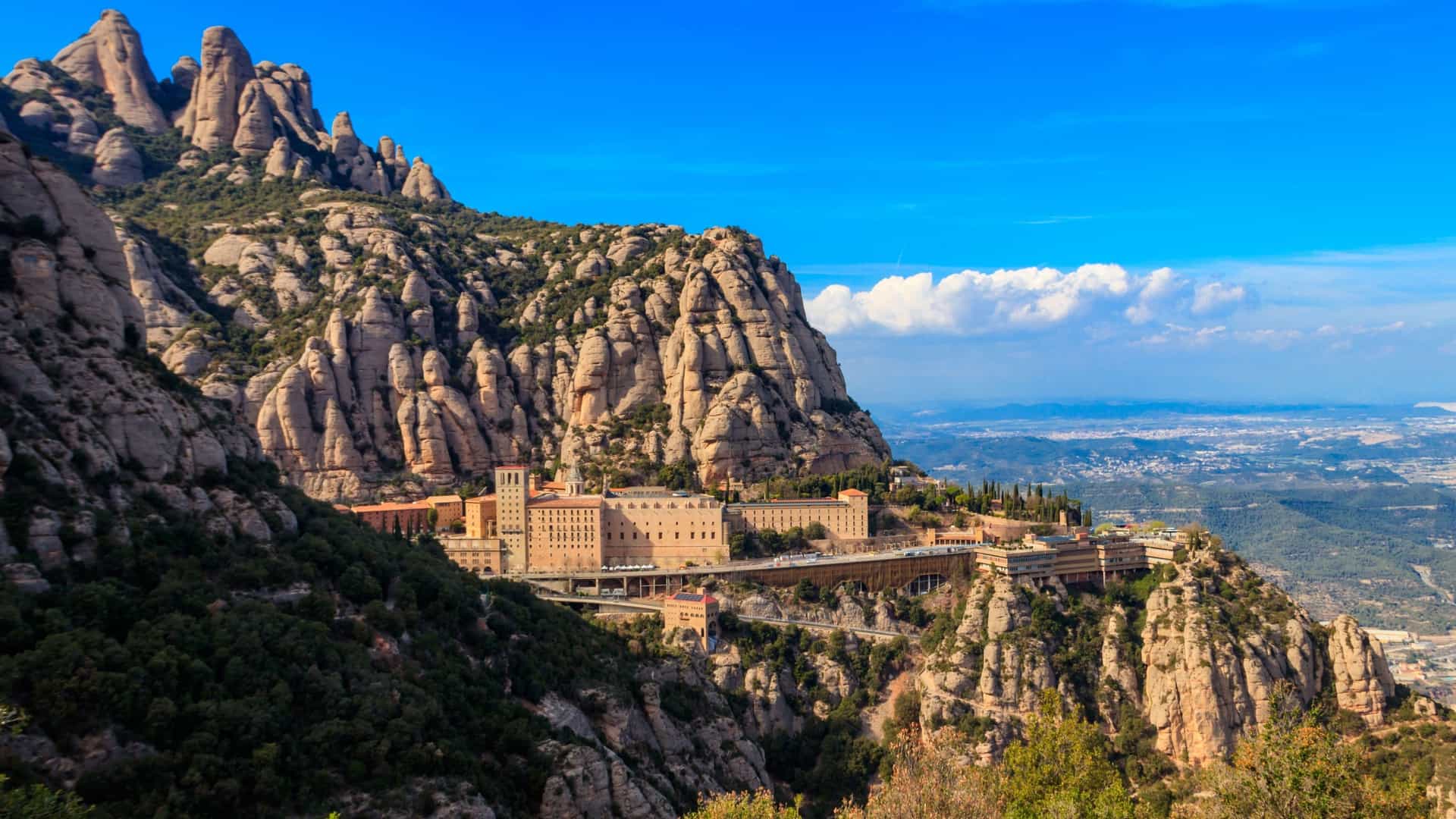 A scenic view of the Santa Maria de Montserrat Abbey, a Benedictine monastery, nestled into the unique, jagged mountain cliffs of Montserrat, Spain, under a bright blue sky.