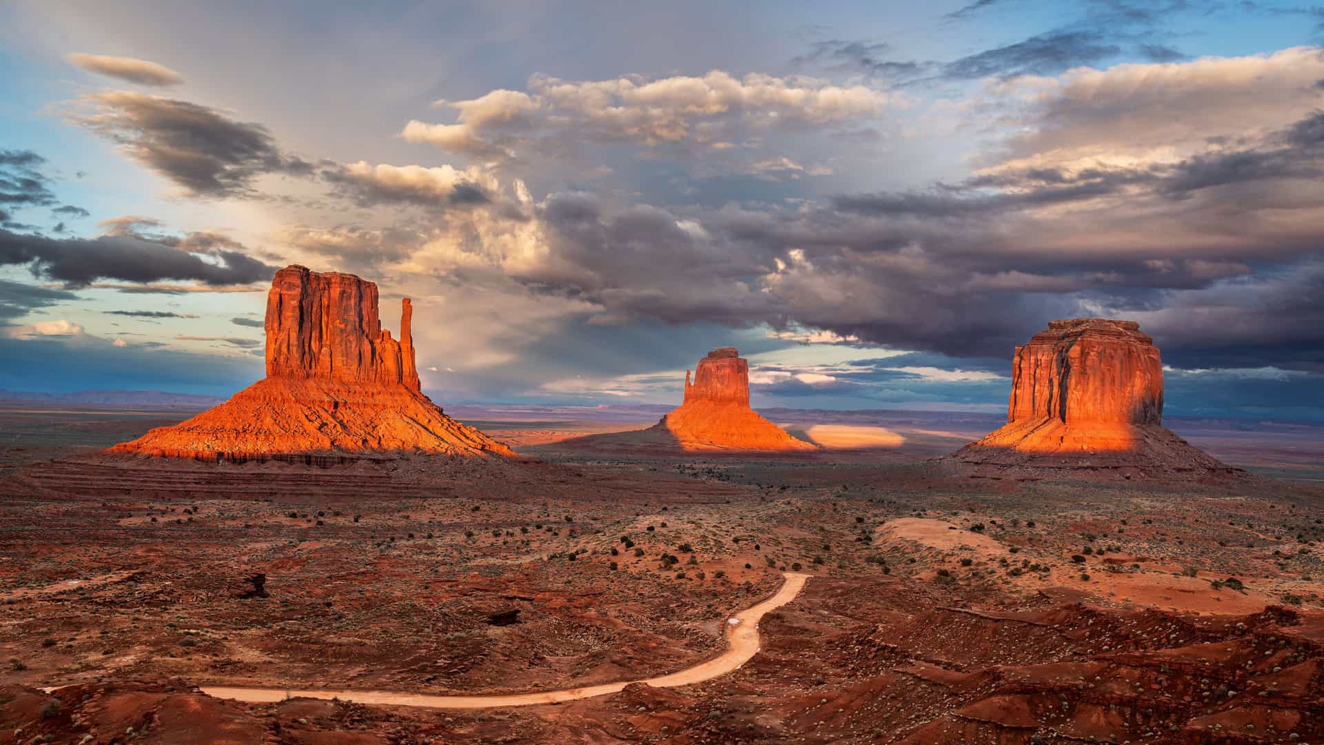 "A dramatic panoramic view of the iconic sandstone buttes and mesas in Monument Valley, Utah, glowing orange at sunset under a moody, cloudy sky.  "