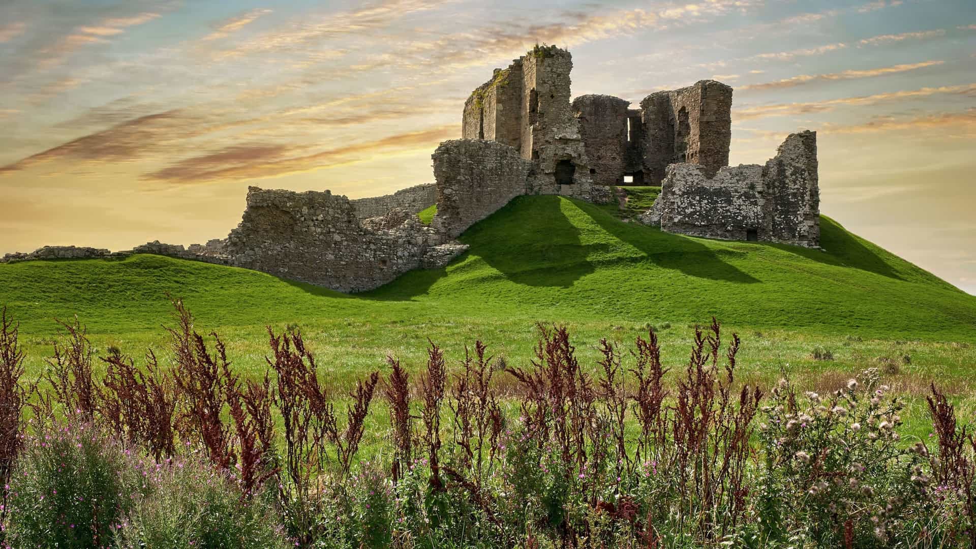 A view of the historic Duffus Castle ruins, a medieval motte-and-bailey fortress in Moray, Scotland, situated on a grassy hill with tall grass and flowers in the foreground.