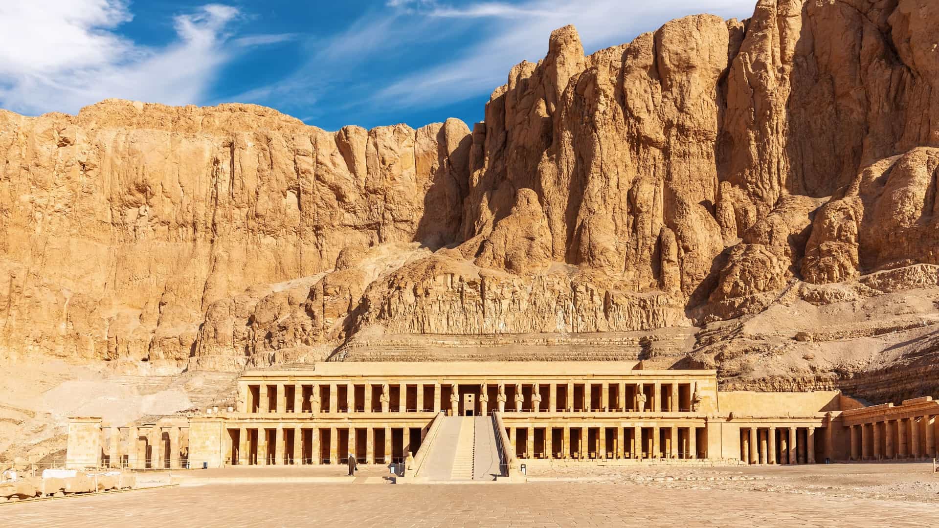 A view of the Mortuary Temple of Hatshepsut, a stunning ancient Egyptian structure with colonnaded terraces, built directly into the side of the towering cliffs of Deir el-Bahri in Luxor.