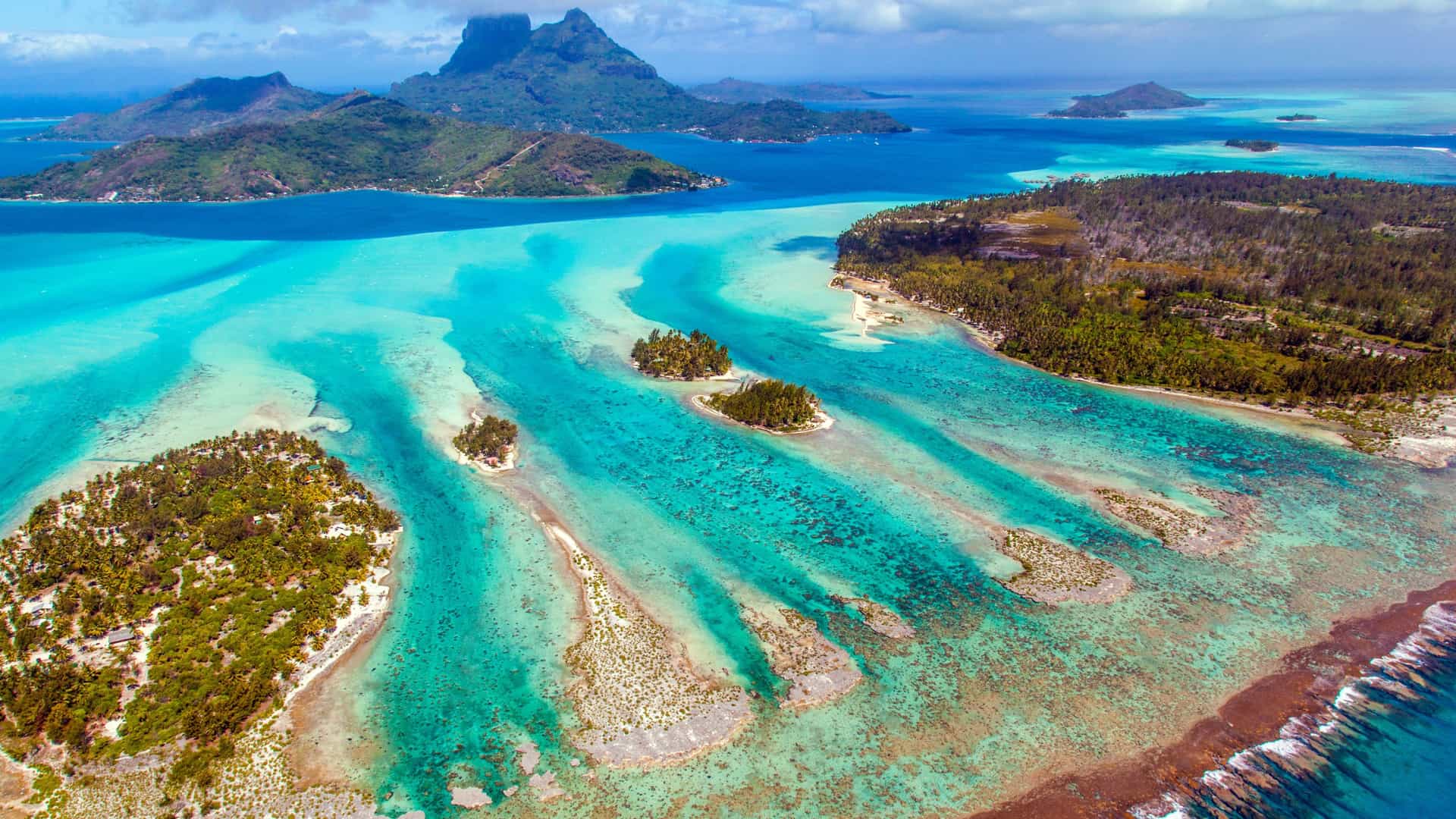 An aerial view of brilliant turquoise lagoons and small motus, with the volcanic peaks of Bora Bora in the background from Motu Tapu.