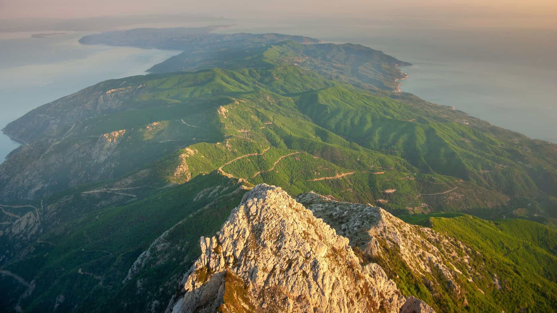 An aerial view of the rugged, green peninsula of Mount Athos in Greece, with its rocky peak rising above the sea and a winding road visible on the hillside.