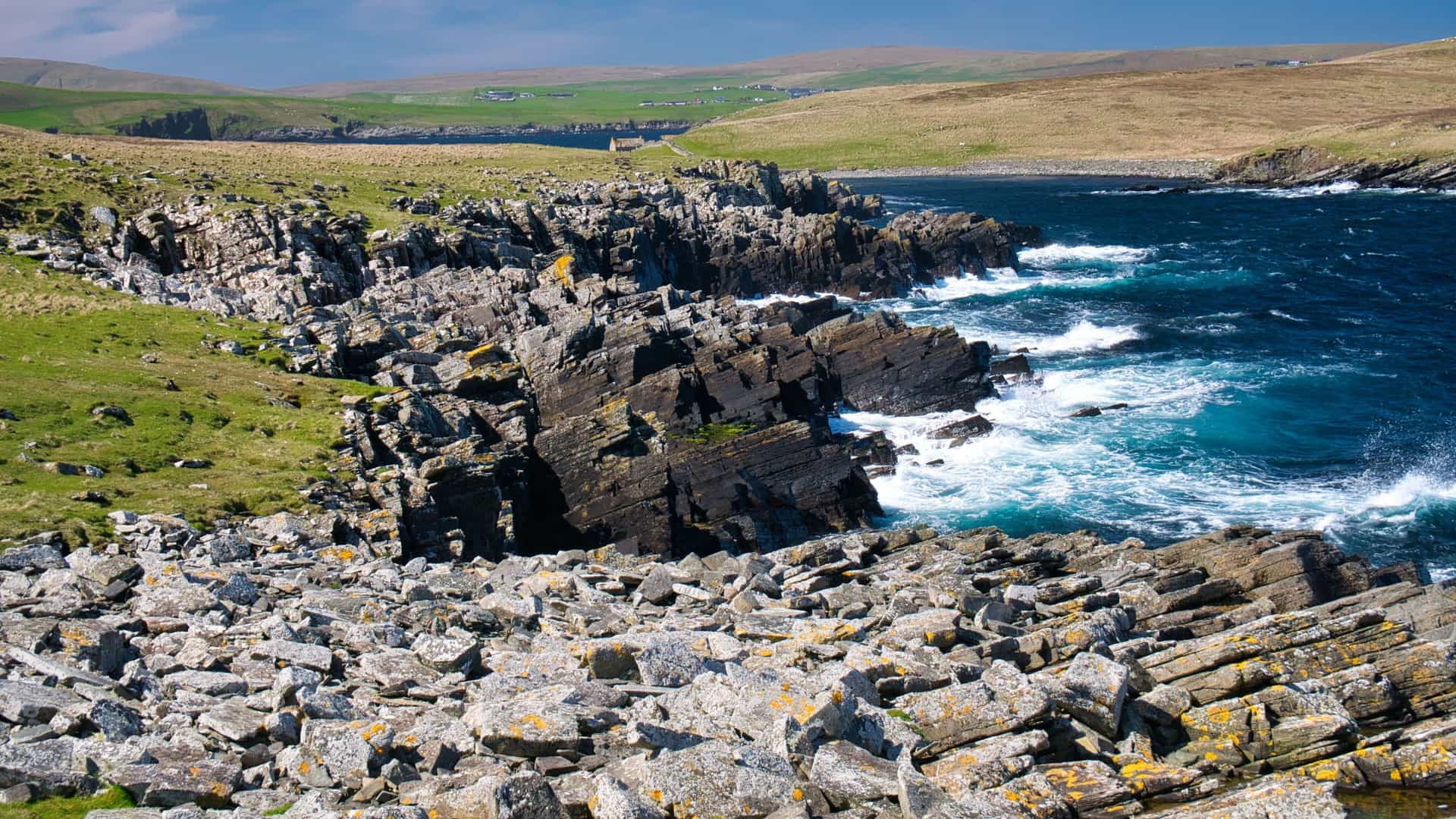 A rocky coastline on the Isle of Mousa in Scotland, with the blue sea crashing against dark cliffs and a rocky foreground under a clear sky.