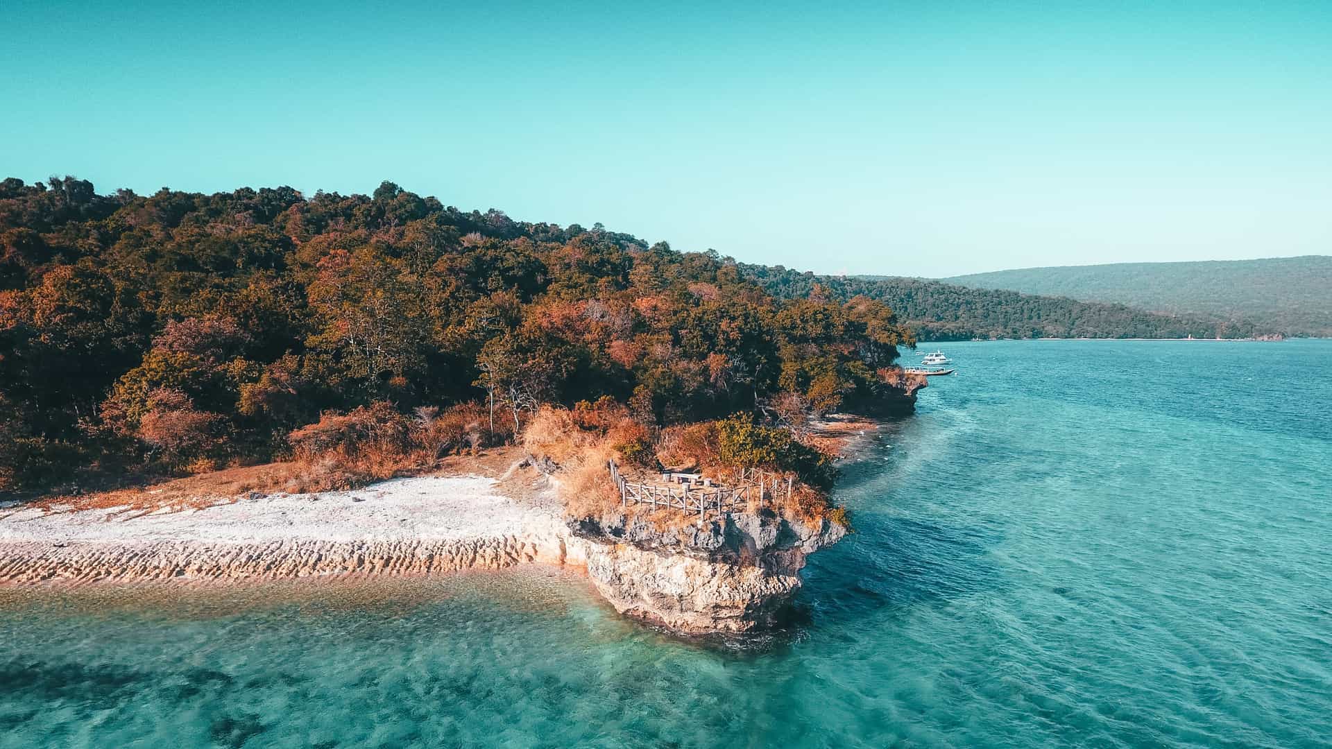 A scenic view of the coastline on Moyo Island, Indonesia, showing a rocky cliff with a wooden fence lookout over crystal-clear turquoise water and lush forested hills.