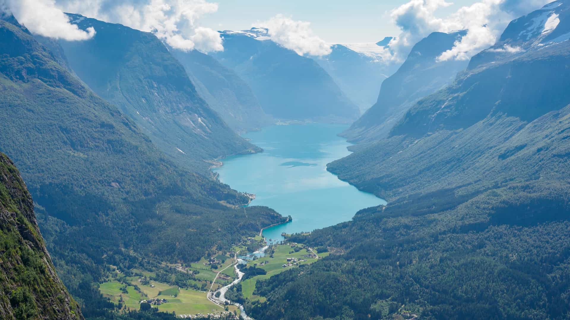 A sweeping fjord landscape in Mt. Hoven with turquoise water winding between steep forested mountains, snow‑touched peaks, scattered shoreline settlements, and clouds drifting along the dramatic valley walls.