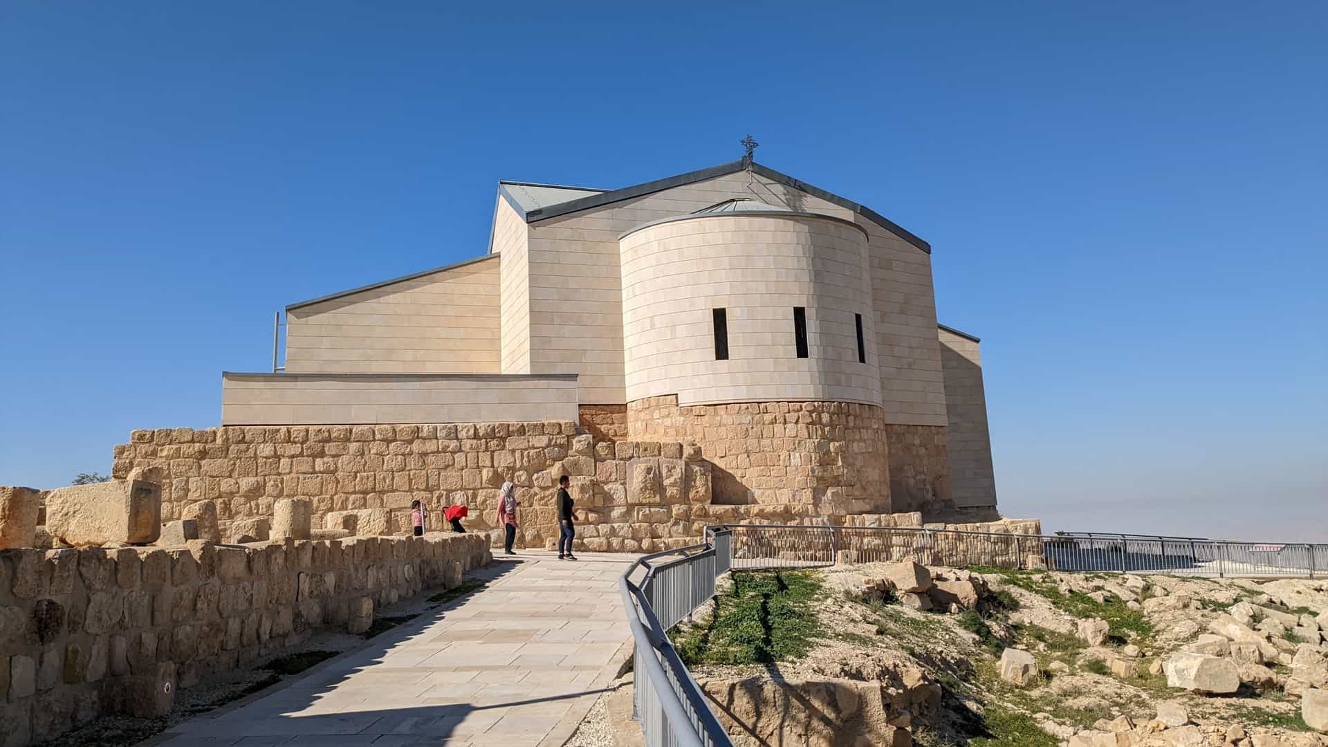 A modern, curved, light-stone sanctuary and church stand on a stone base on Mount Nebo, Jordan, under a bright blue sky.