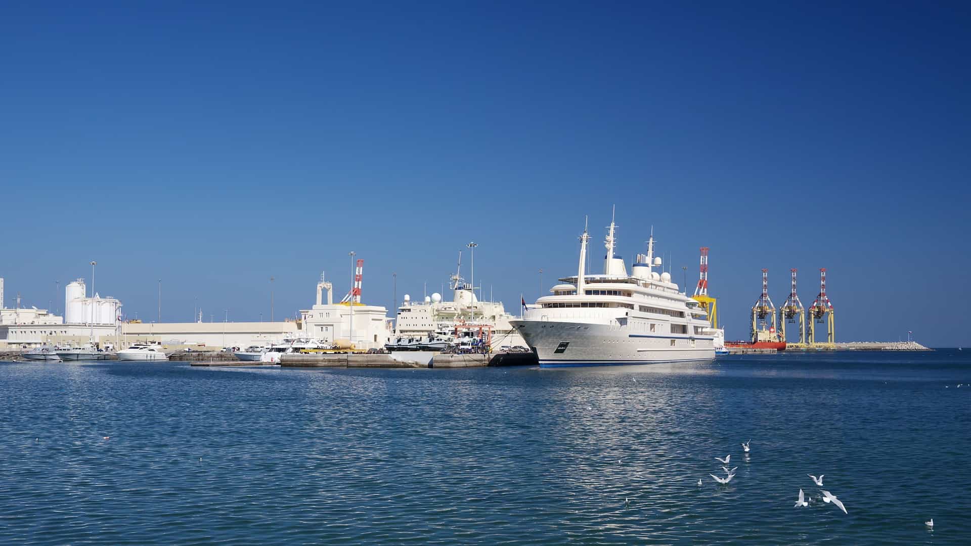 A high-angle view of Port Sultan Qaboos in Muscat, Oman, with a large cruise ship or cargo vessel docked in the harbor. The port is backed by the residential buildings of the city's waterfront, which climb the surrounding rugged, arid mountains. The scene captures the commercial and scenic beauty of Muscat's main port.