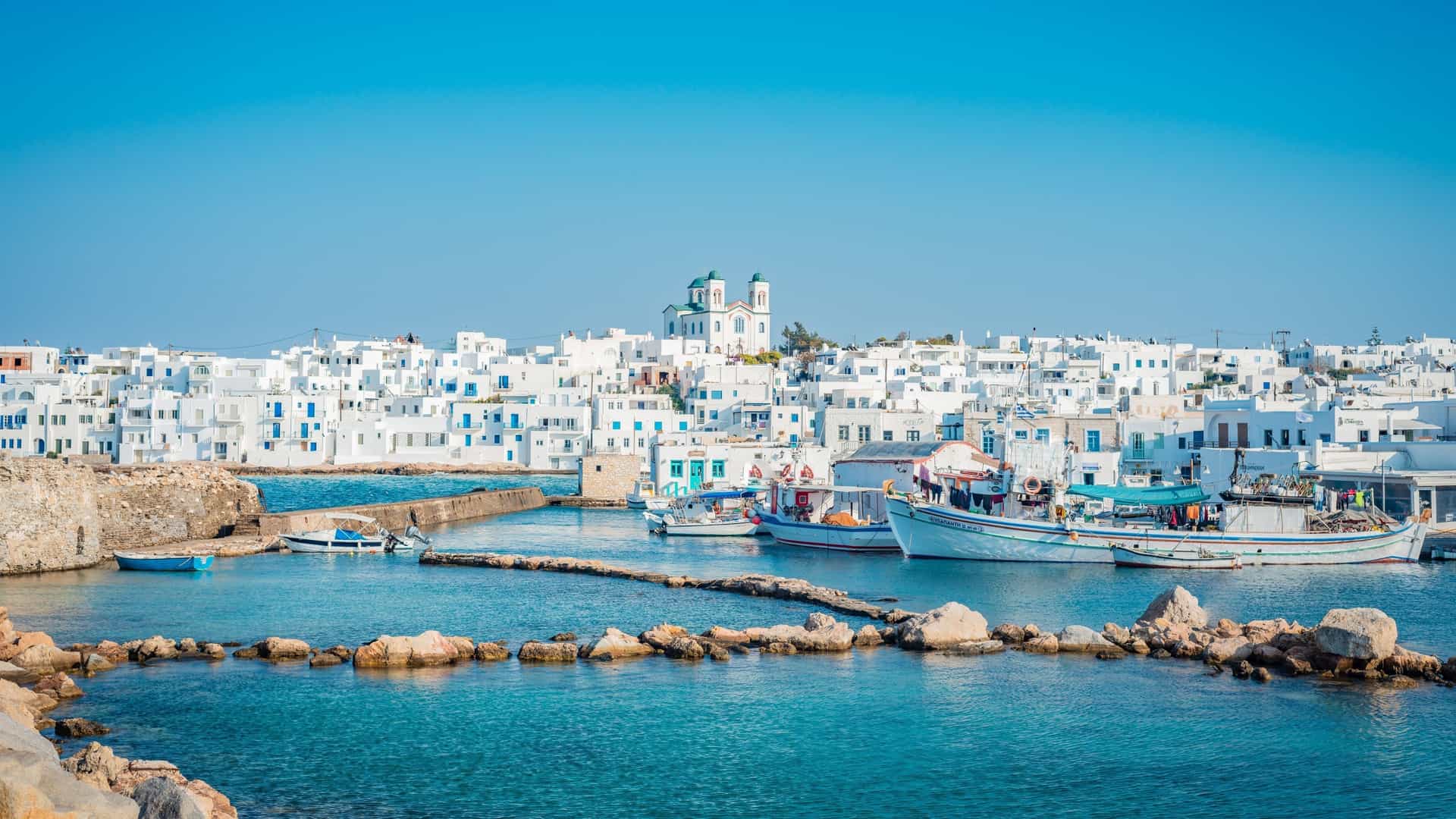 The harbor of Naousa, Greece, featuring white Cycladic buildings, a dome church, and fishing boats in the clear blue water.