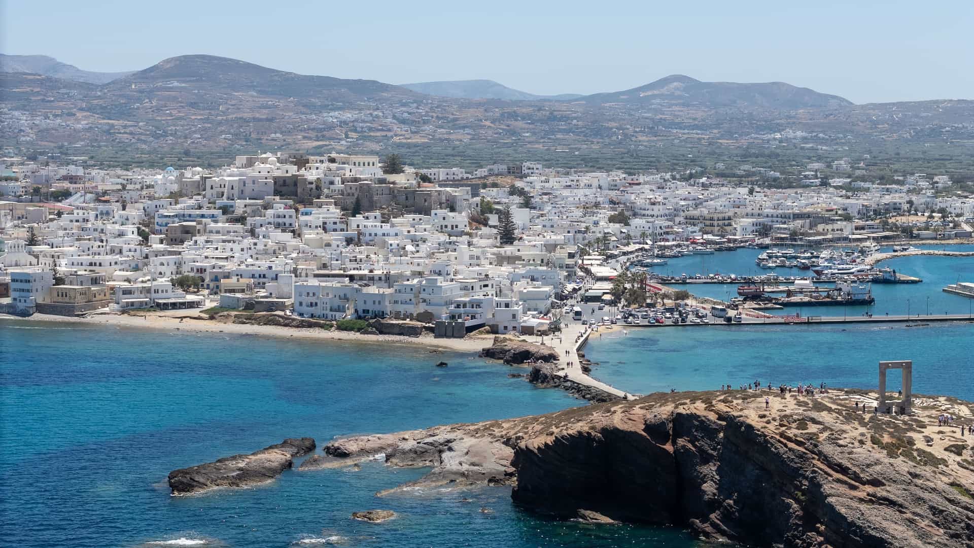 A scenic aerial view of Naxos Town, Greece, with its traditional white buildings, a bustling harbor, and the ancient Portara temple gate on a rocky peninsula.