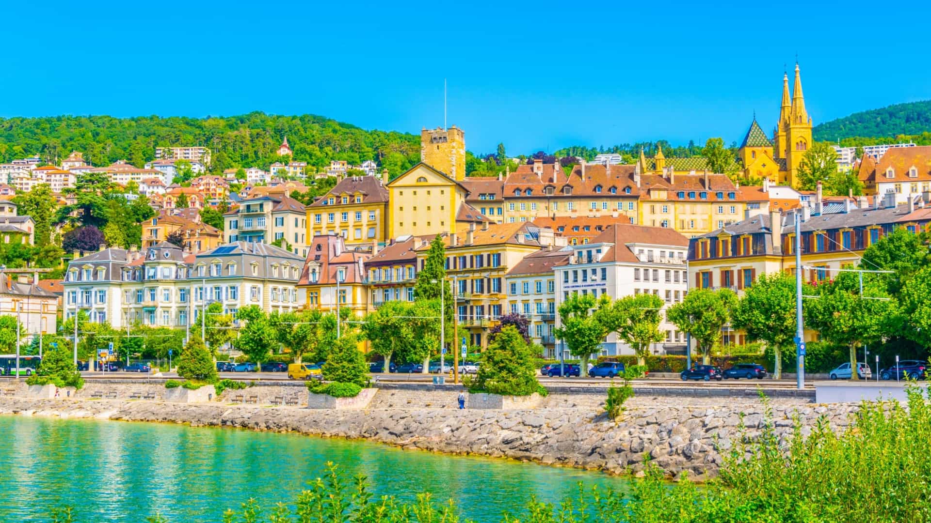 A scenic view of the charming town of Neuchâtel, Switzerland, with its colorful historic buildings and a castle perched on a hillside overlooking a tranquil lake.
