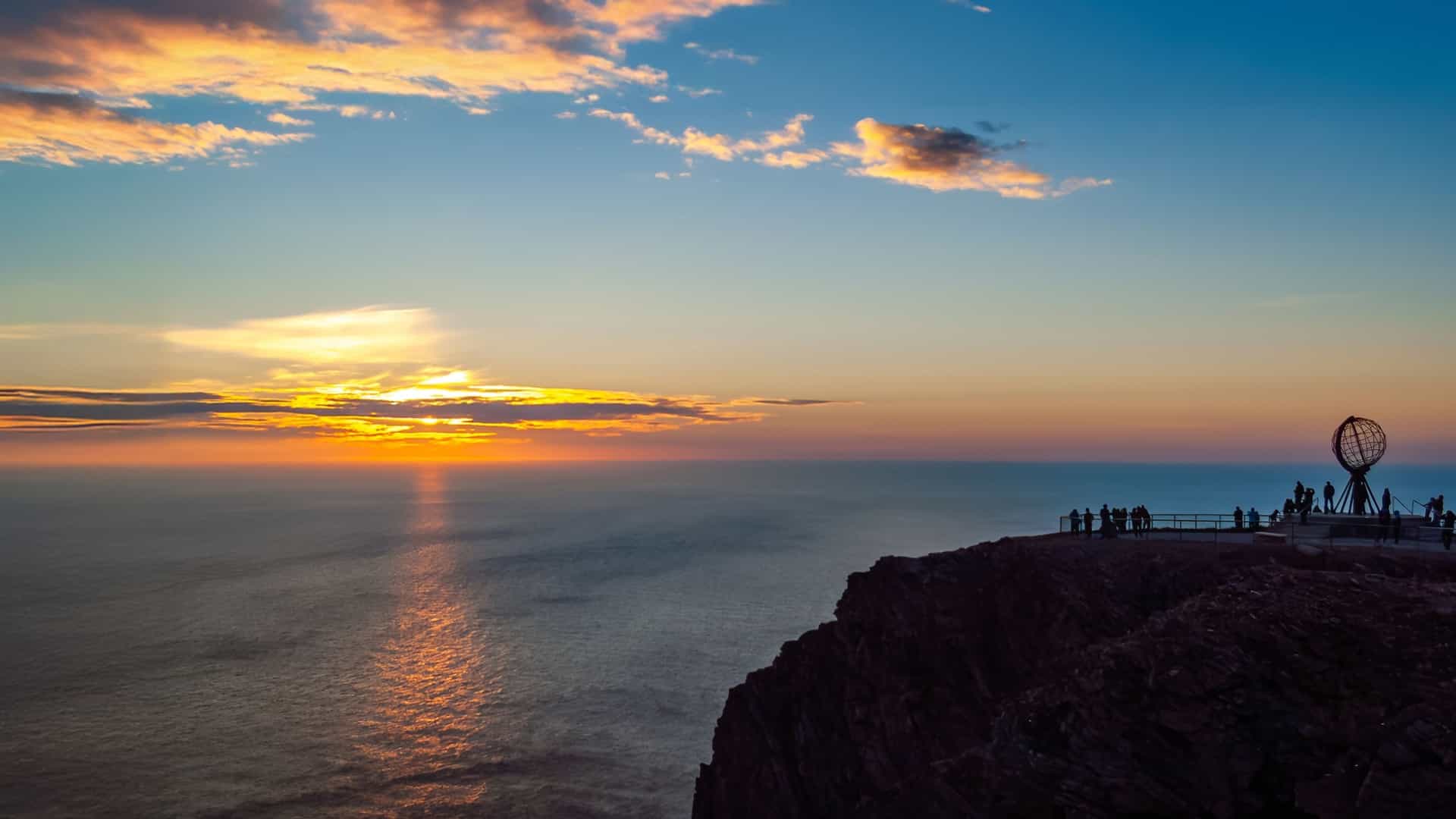 A dramatic Arctic scene in Nordkapp with a glowing orange‑and‑blue horizon over the ocean, a dark cliff edge, and visitors gathered beside the iconic globe monument silhouetted against the vivid sky.