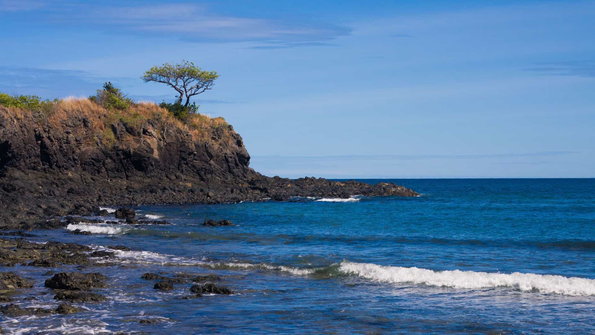 A rocky shoreline on the island of Nosy Be, Madagascar, with waves crashing against the black volcanic rock, a lone tree on the cliffside, and the vast Indian Ocean under a blue sky.