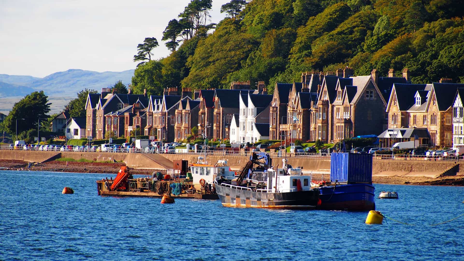 A vibrant shot of the waterfront in Oban, Scotland, with fishing boats docked in the bay in front of a row of Victorian-era townhouses and a lush green hillside.