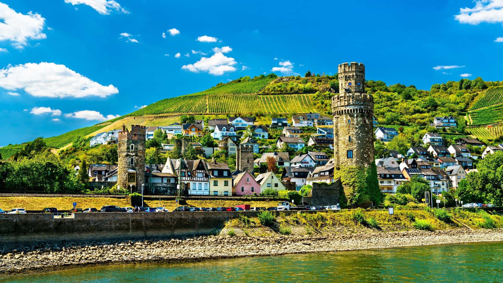 A colorful view of Oberwesel, Germany, showing a row of medieval houses and two stone towers along the Rhine River with steep, terraced vineyards in the background.