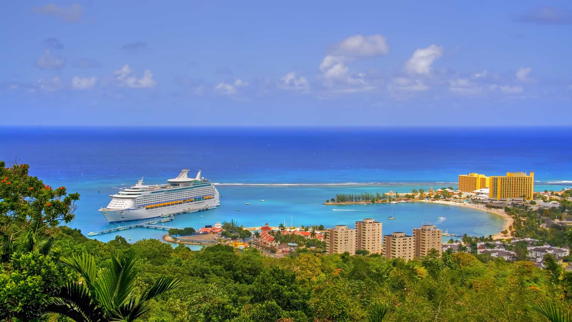 An elevated panoramic shot of the beautiful Ocho Rios Bay in Jamaica, showing a large cruise ship docked at the pier, a sandy beach, and luxury resorts under a blue sky.
