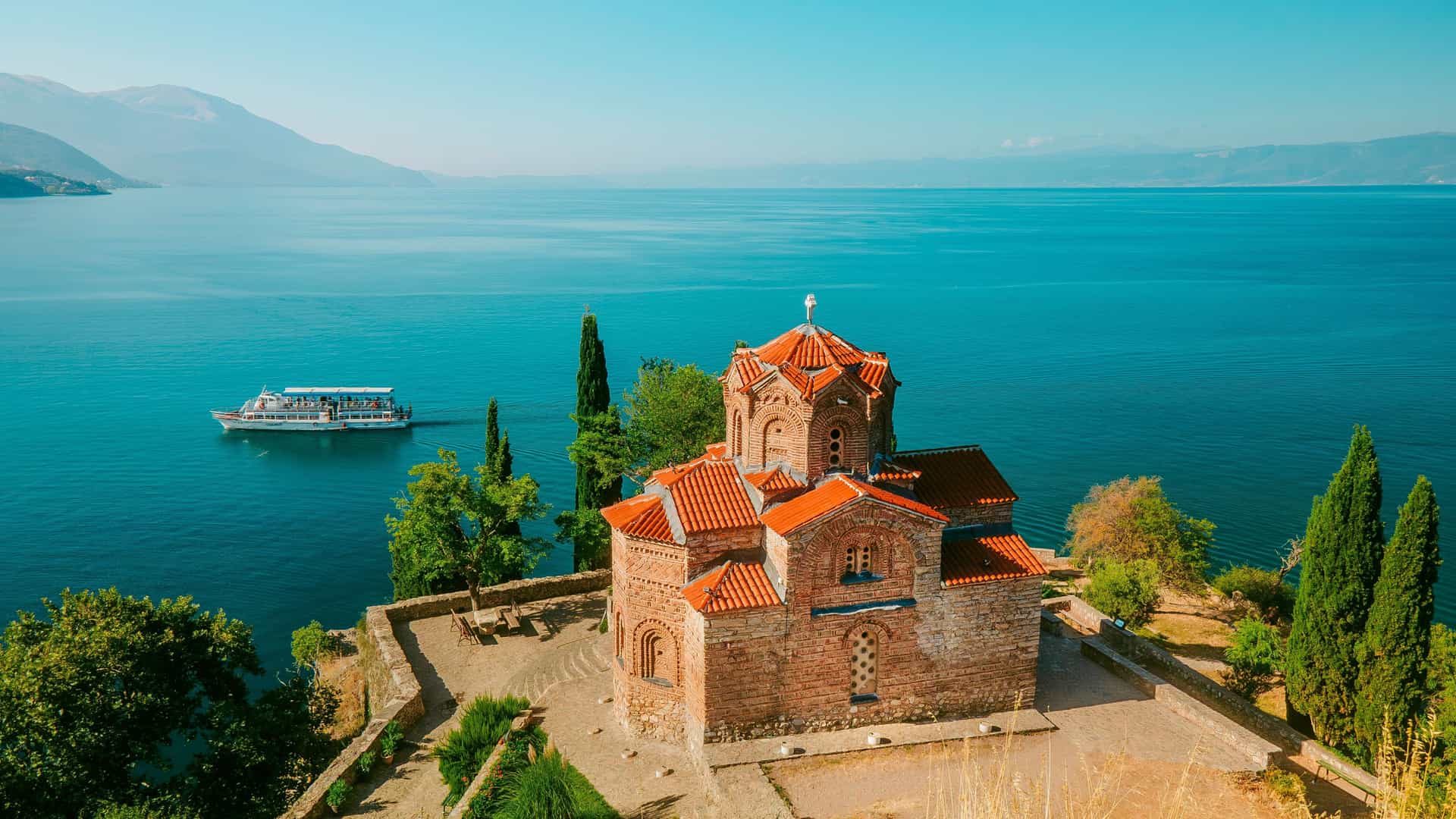 "A panoramic view of the historic Church of St. John the Theologian at Kaneo, overlooking the serene blue waters of Lake Ohrid in Ohrid, North Macedonia, with a tour boat in the background.  "