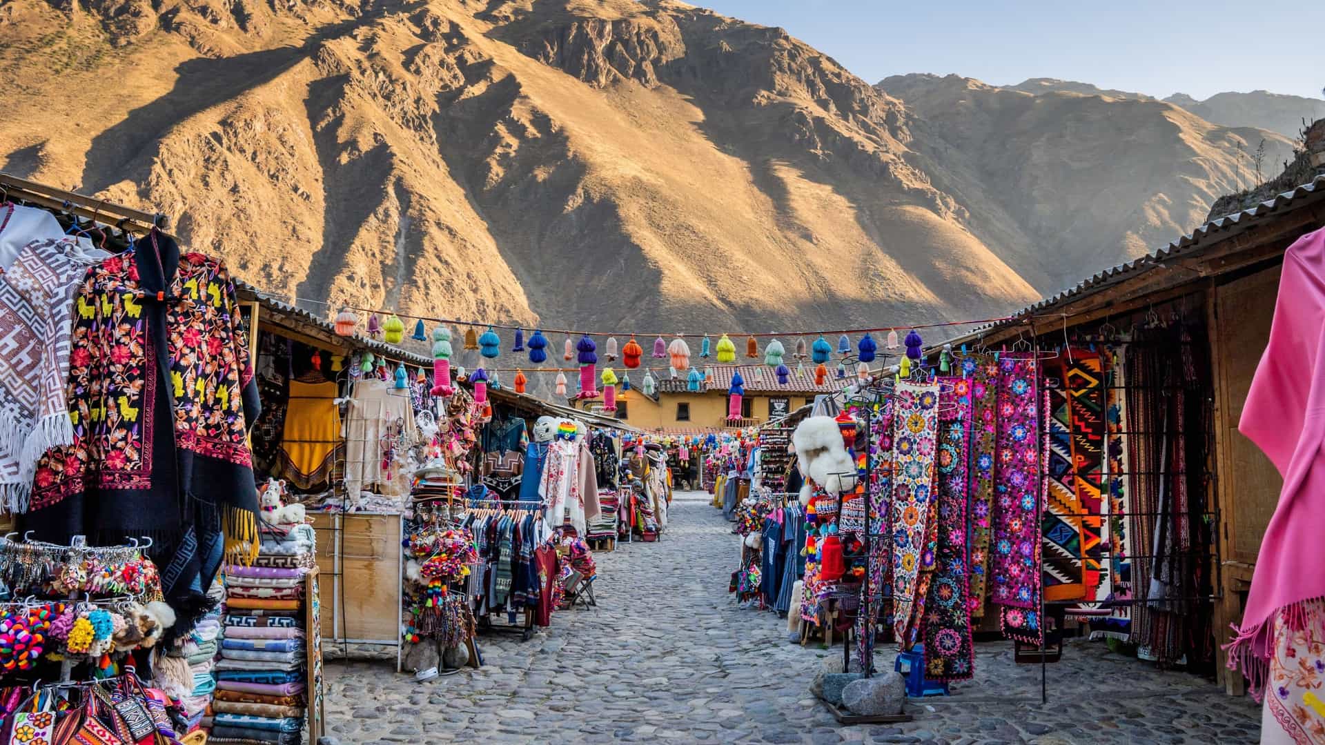 A lively shot of a colorful market street in Ollantaytambo, Peru, with stalls selling traditional textiles and goods, set against a backdrop of the Andes mountains.