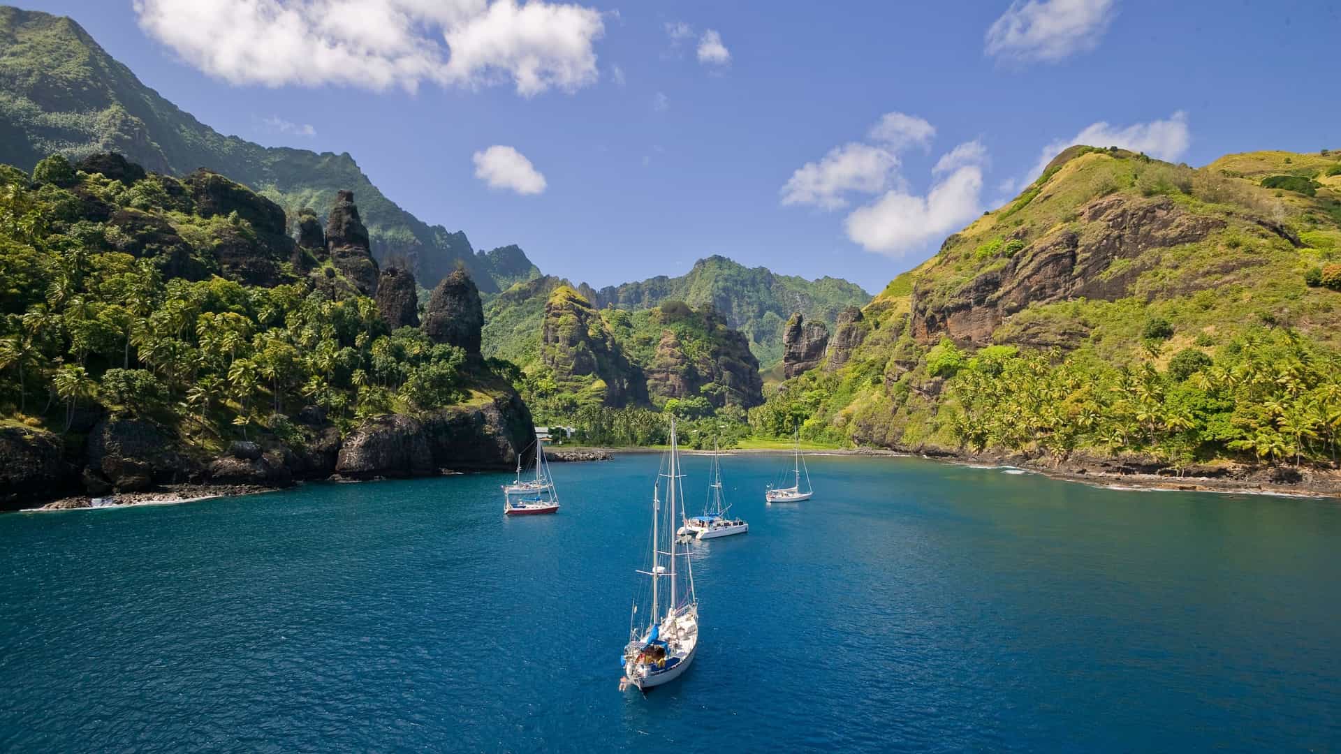 A beautiful scenic view of Omoa Bay on Fatu Hiva, Marquesas Islands, with several sailboats anchored in the peaceful blue water, surrounded by dramatic green cliffs and lush tropical vegetation.