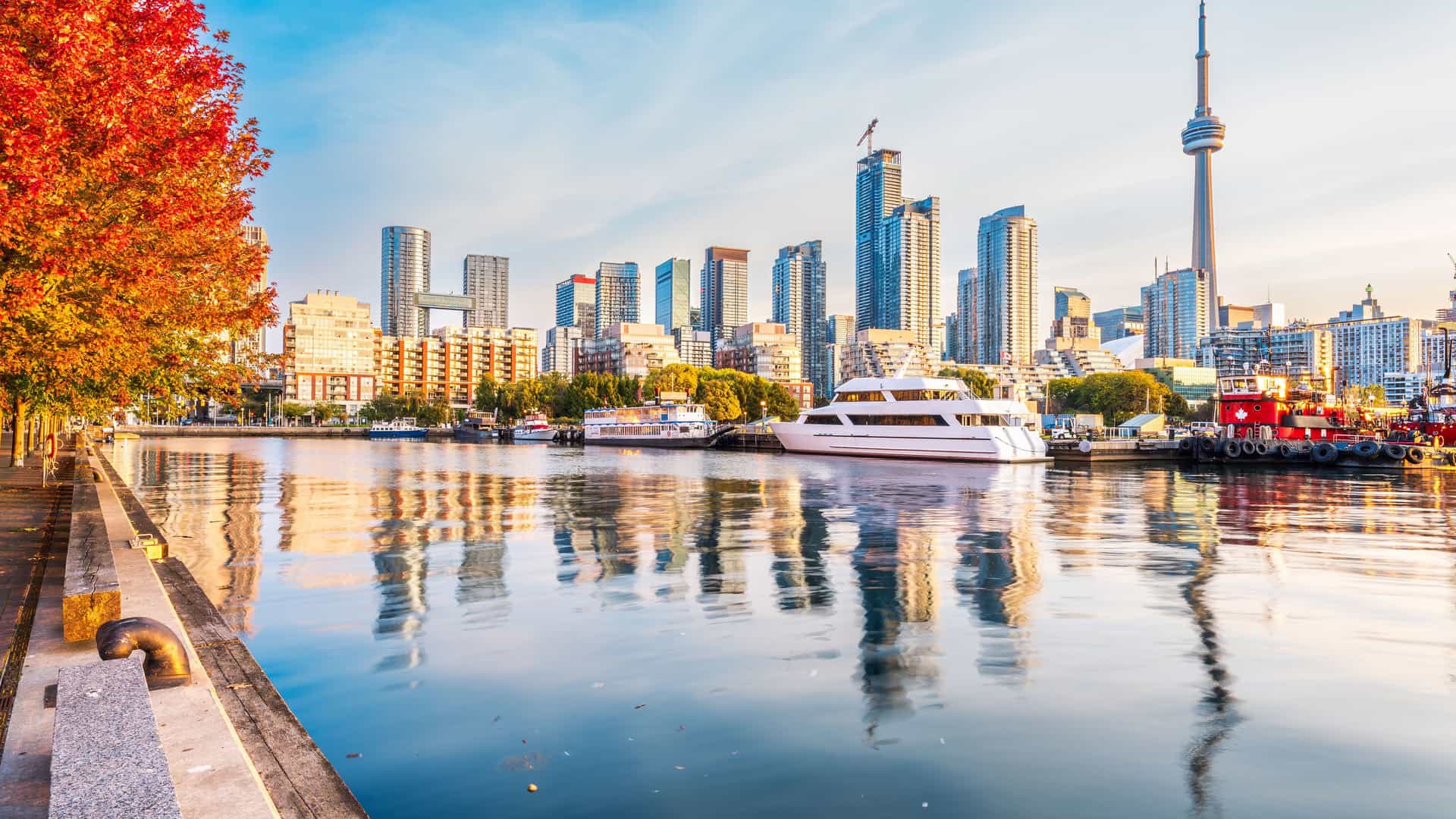 "A vibrant autumn panoramic view of the Toronto skyline at sunrise, showing the CN Tower, yachts, and the cityscape reflecting off the calm waters of Lake Ontario.  "